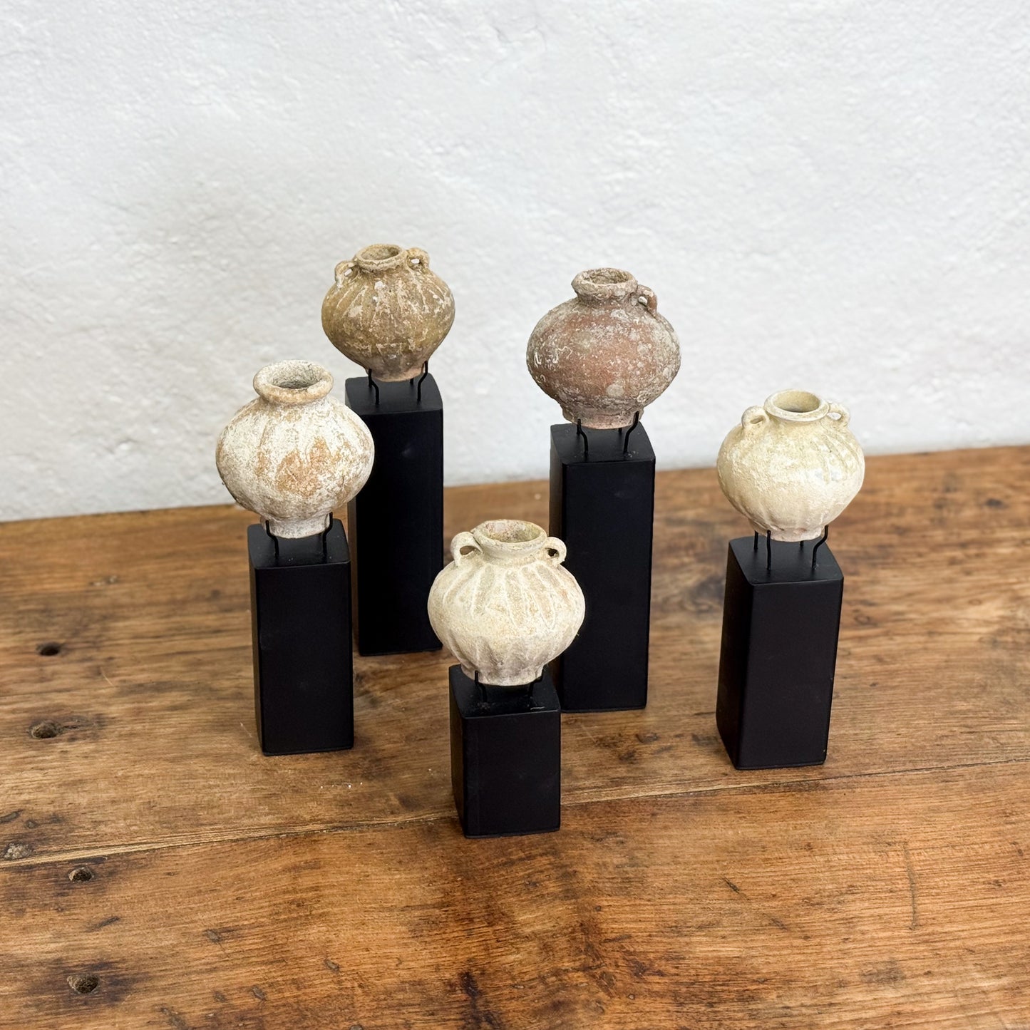 Set of six ceramic vases on black stands against a white textured wall.