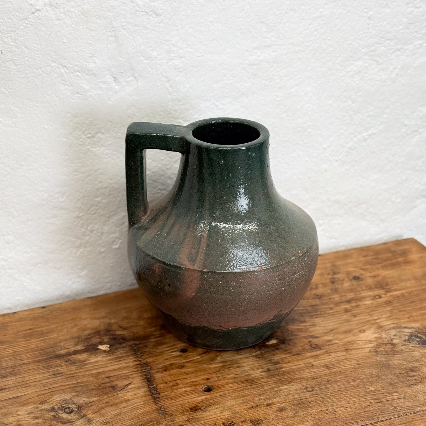 Green ceramic pitcher on a wooden surface with a white textured wall background