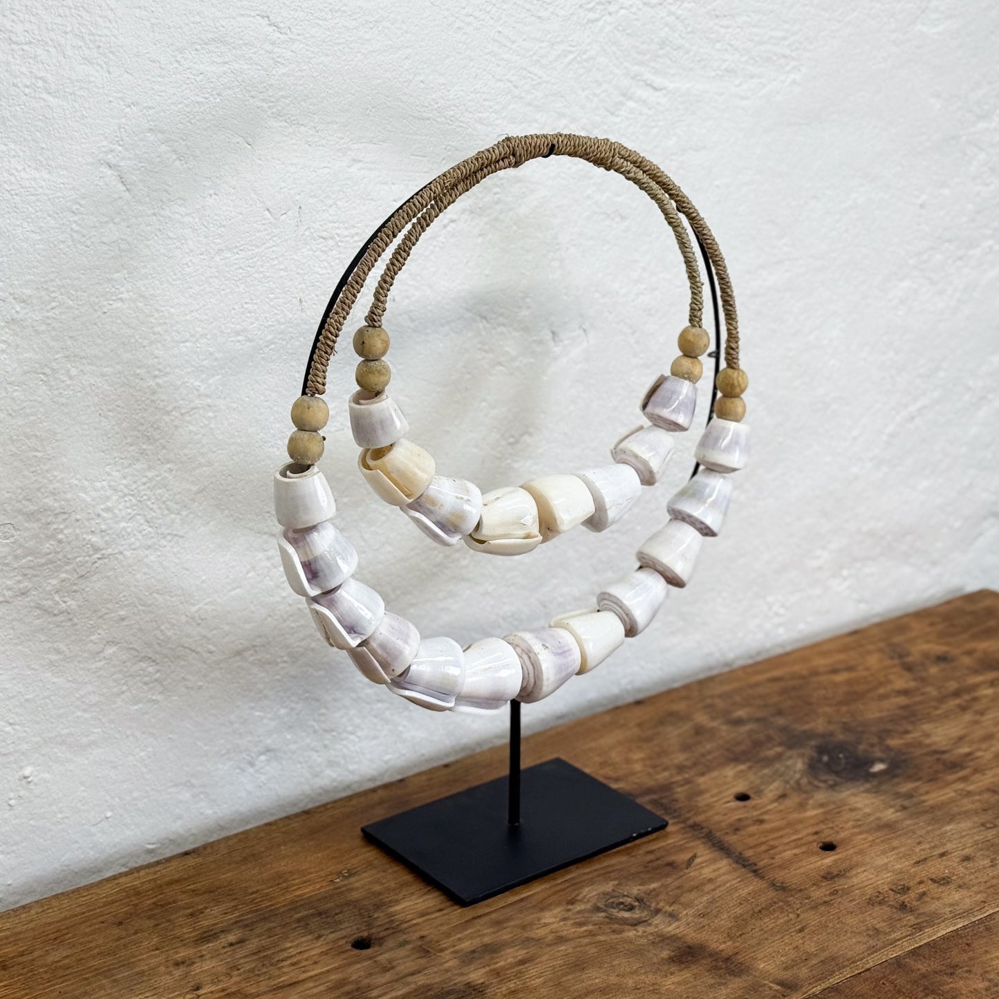 Decorative ring with white beads and brown rope on a black stand against a white wall.