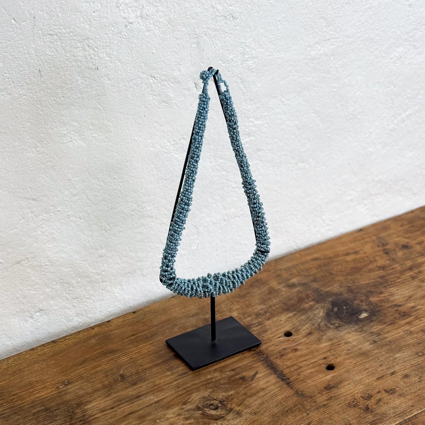 Blue woven necklace on a black stand against a white wall.