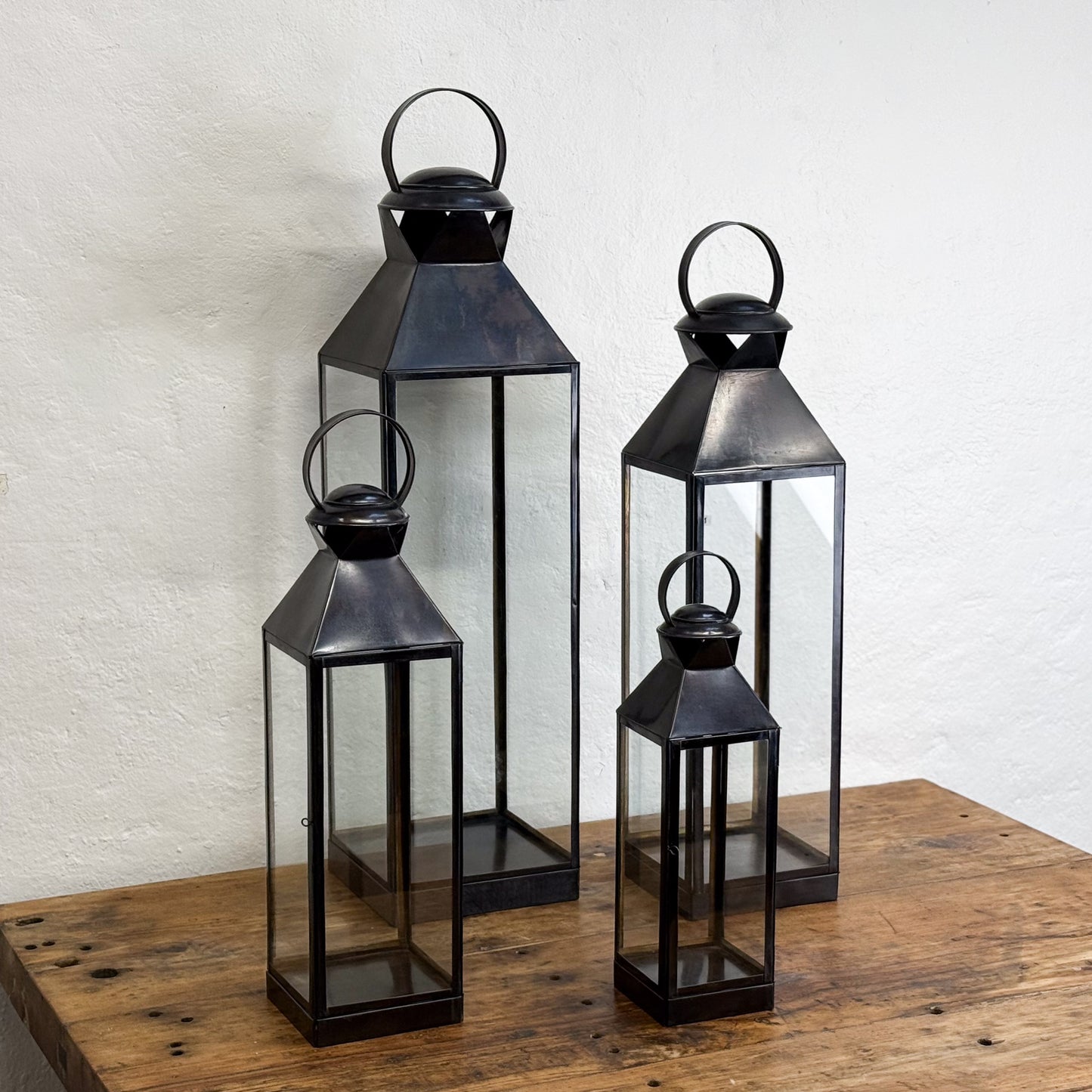 Set of four black metal lanterns on a wooden surface with a white textured wall background.