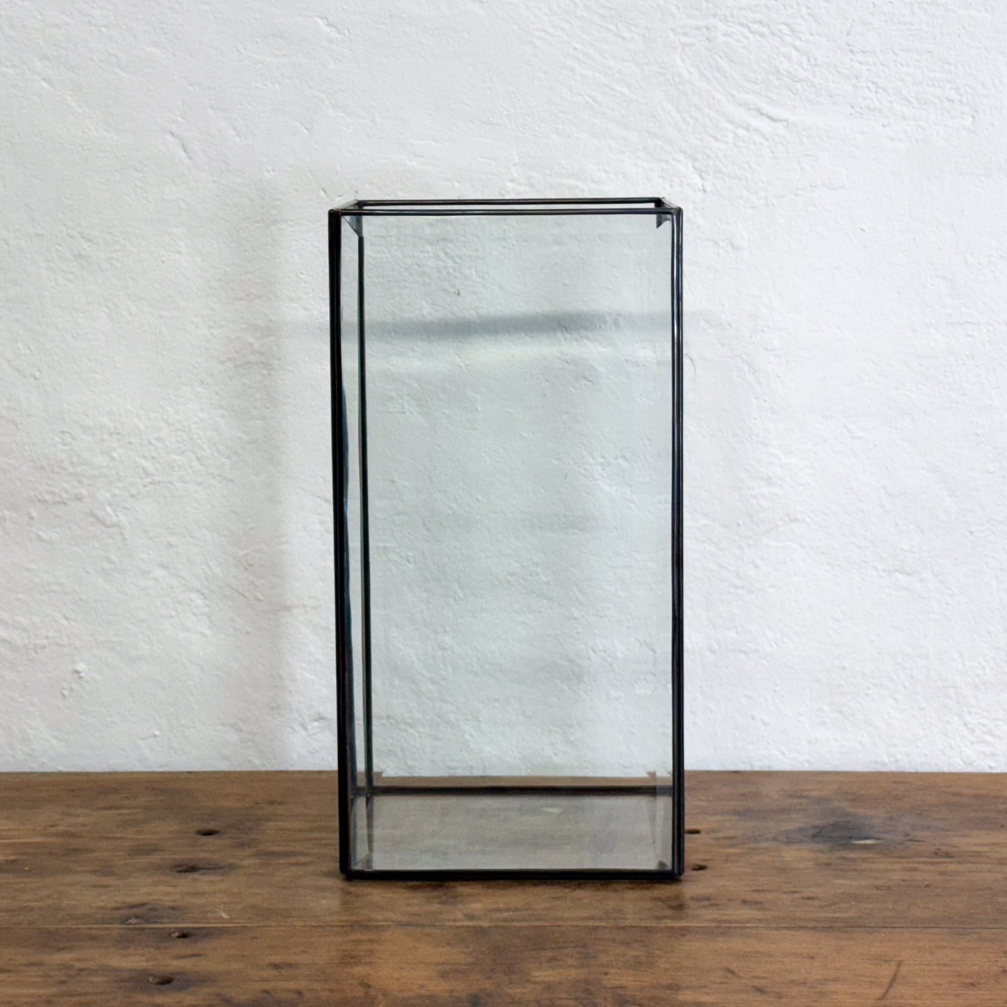 Glass display case with black metal frame on a wooden surface and white textured wall background