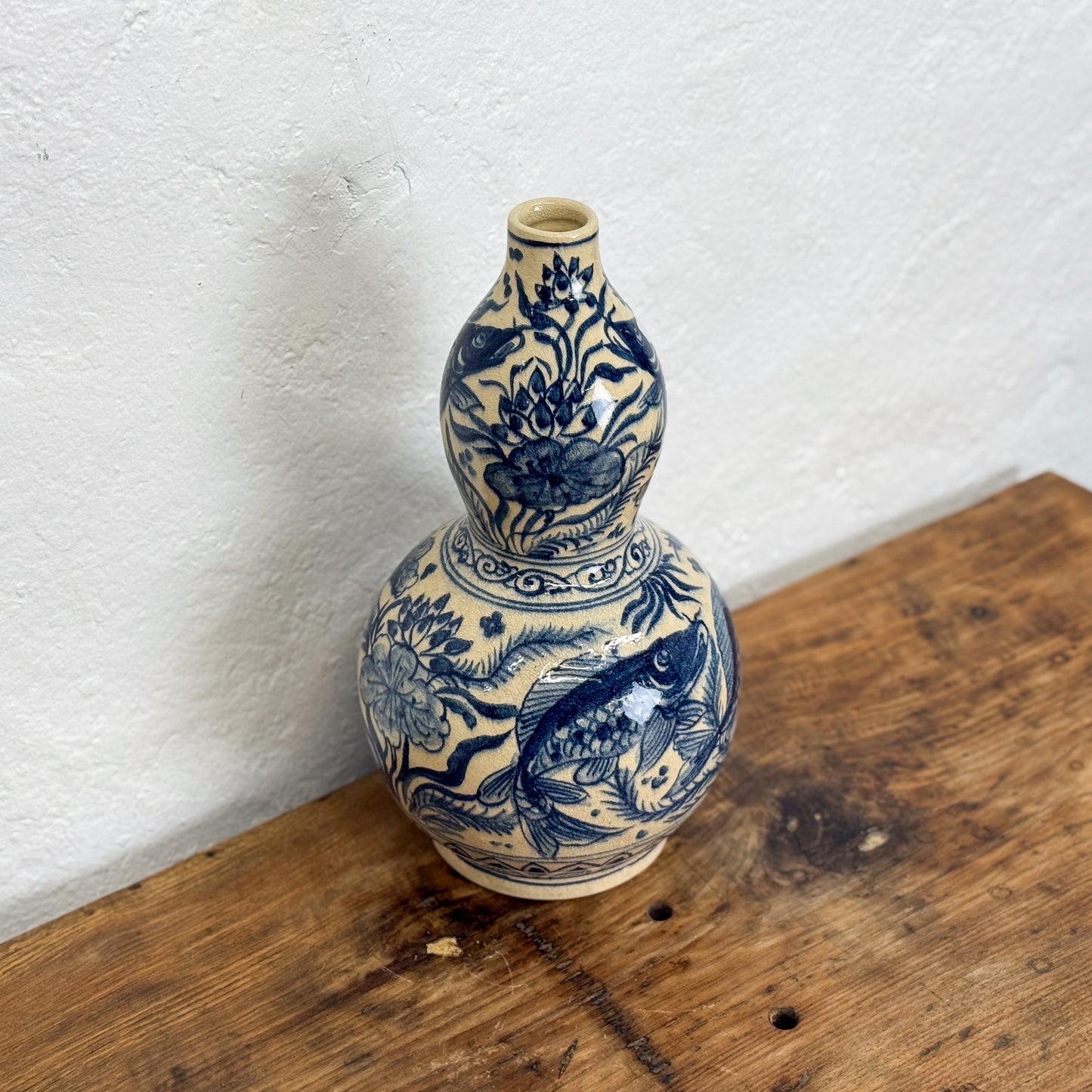 Blue and white ceramic vase with floral and fish design on a wooden surface against a white wall.