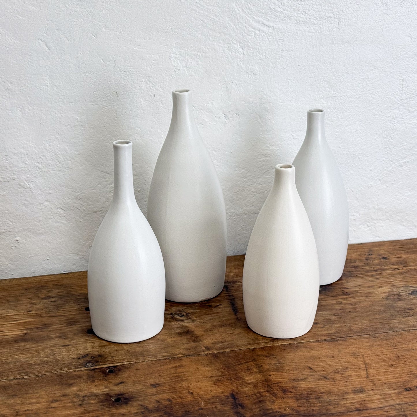 Four white ceramic vases of varying sizes on a wooden surface with a white wall background.