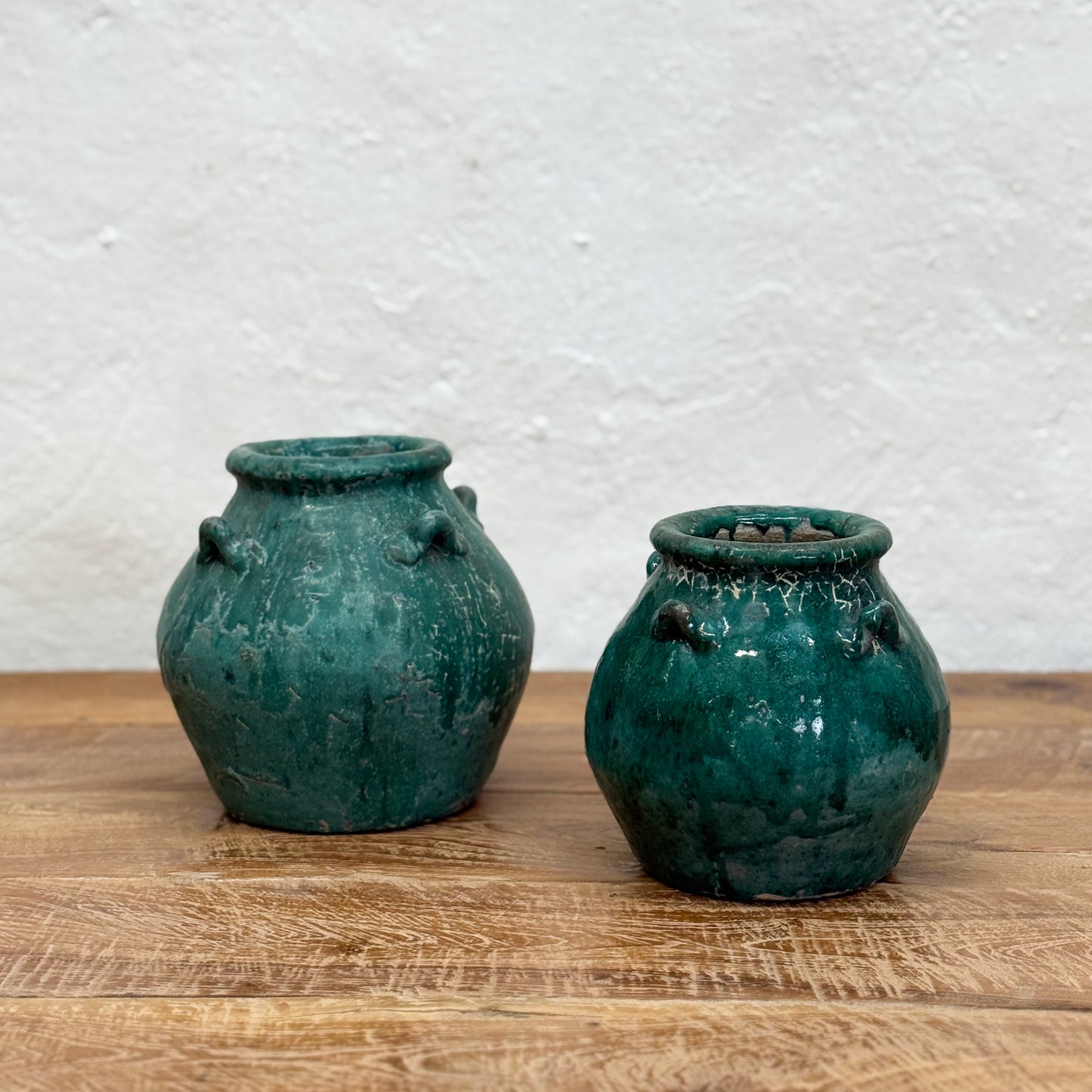 Two green ceramic pots on a wooden surface with a textured white wall background