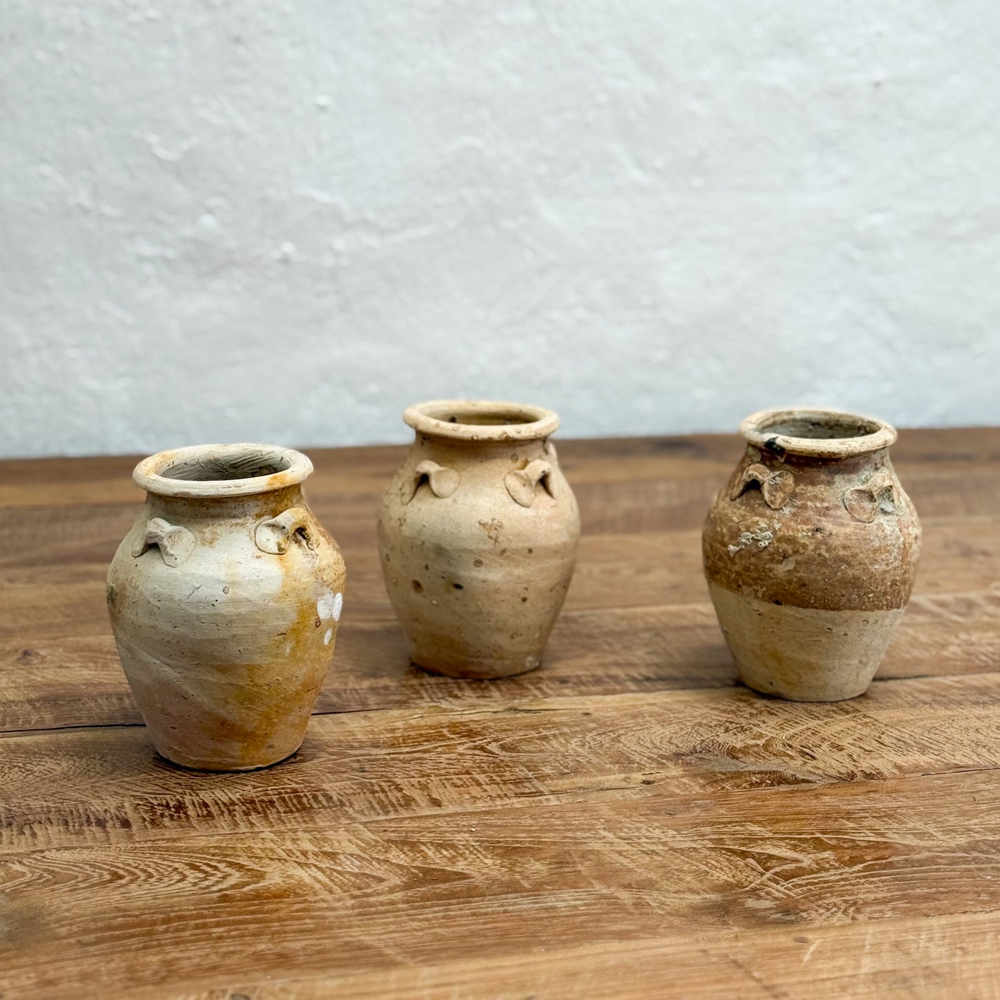 Three ceramic jars with handles on a wooden surface against a textured wall.