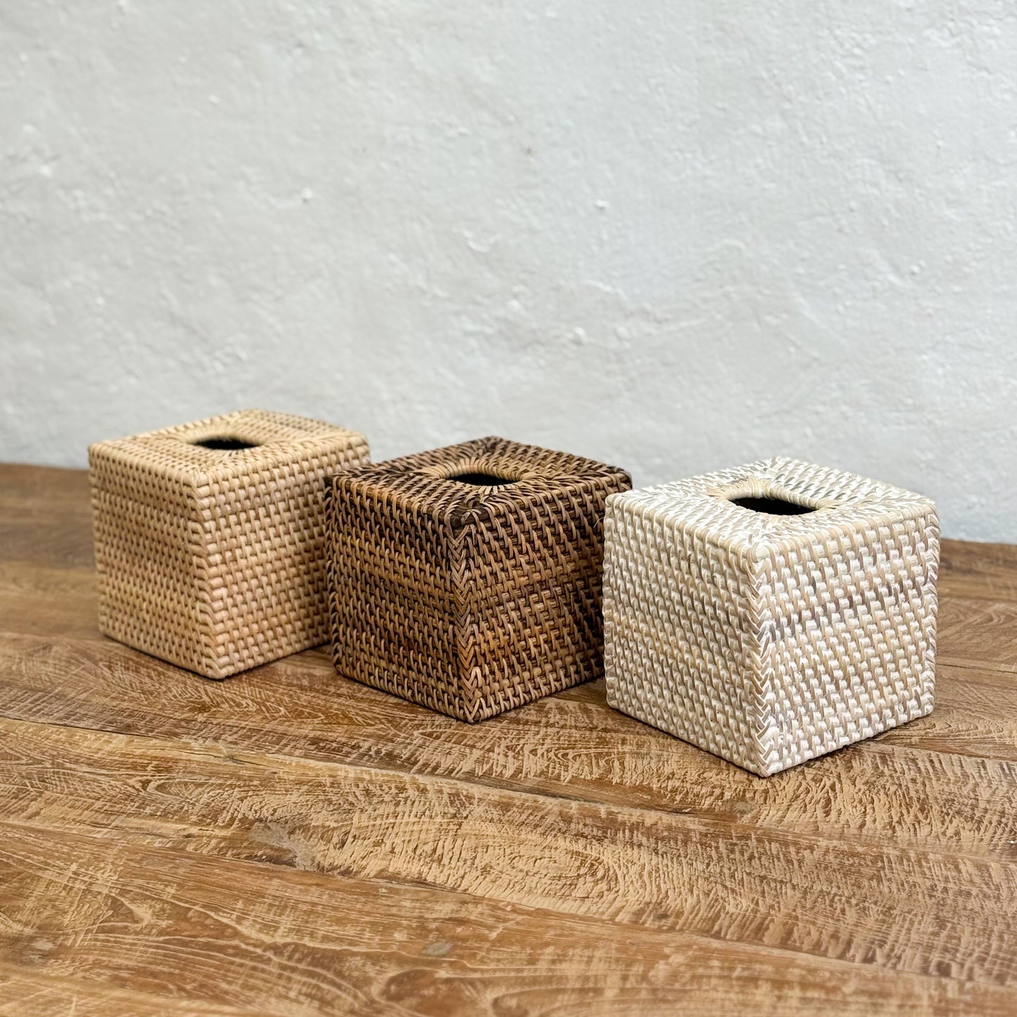 Three woven tissue boxes in different colors on a wooden surface with a gray textured wall background.
