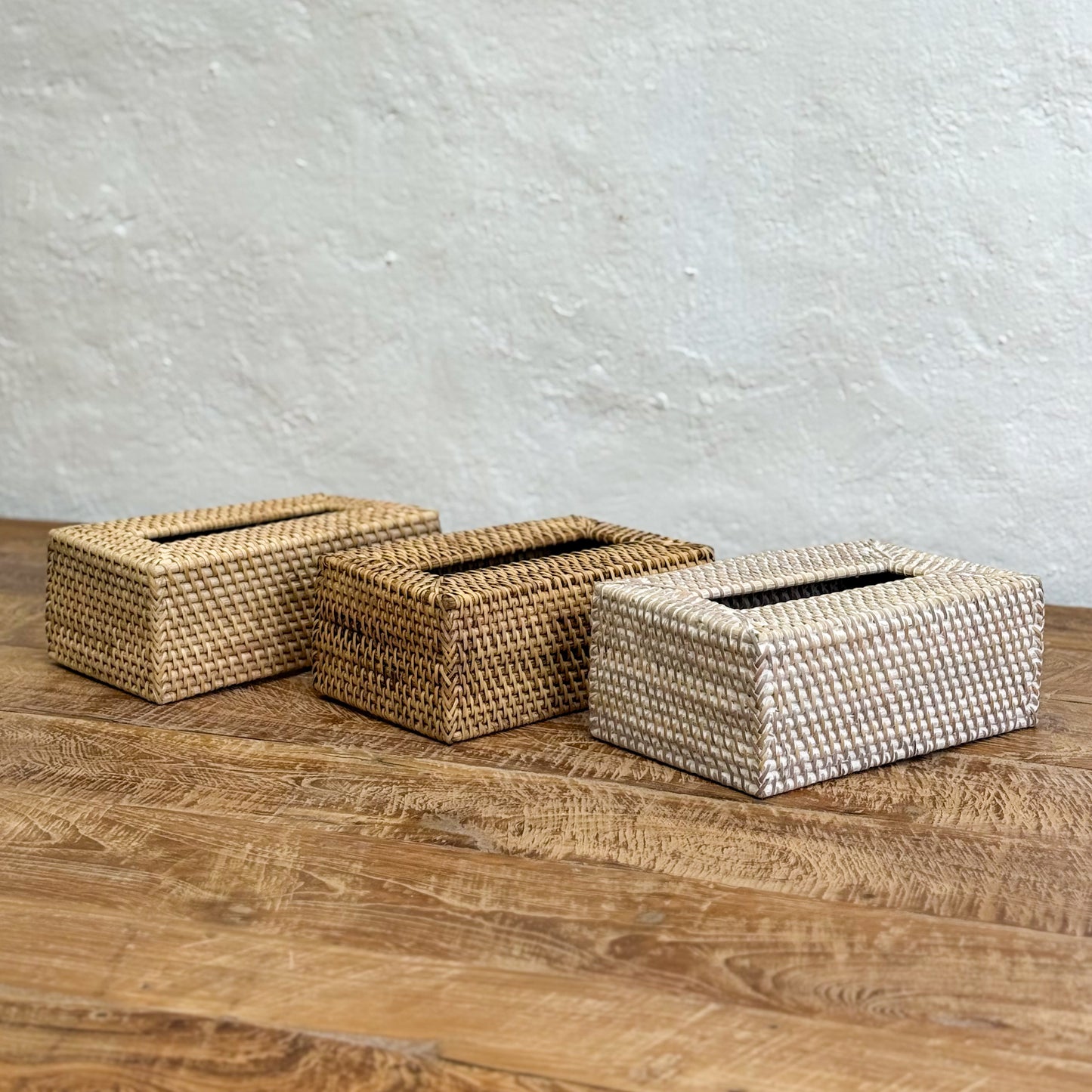 Three woven tissue boxes on a wooden surface with a textured white wall background