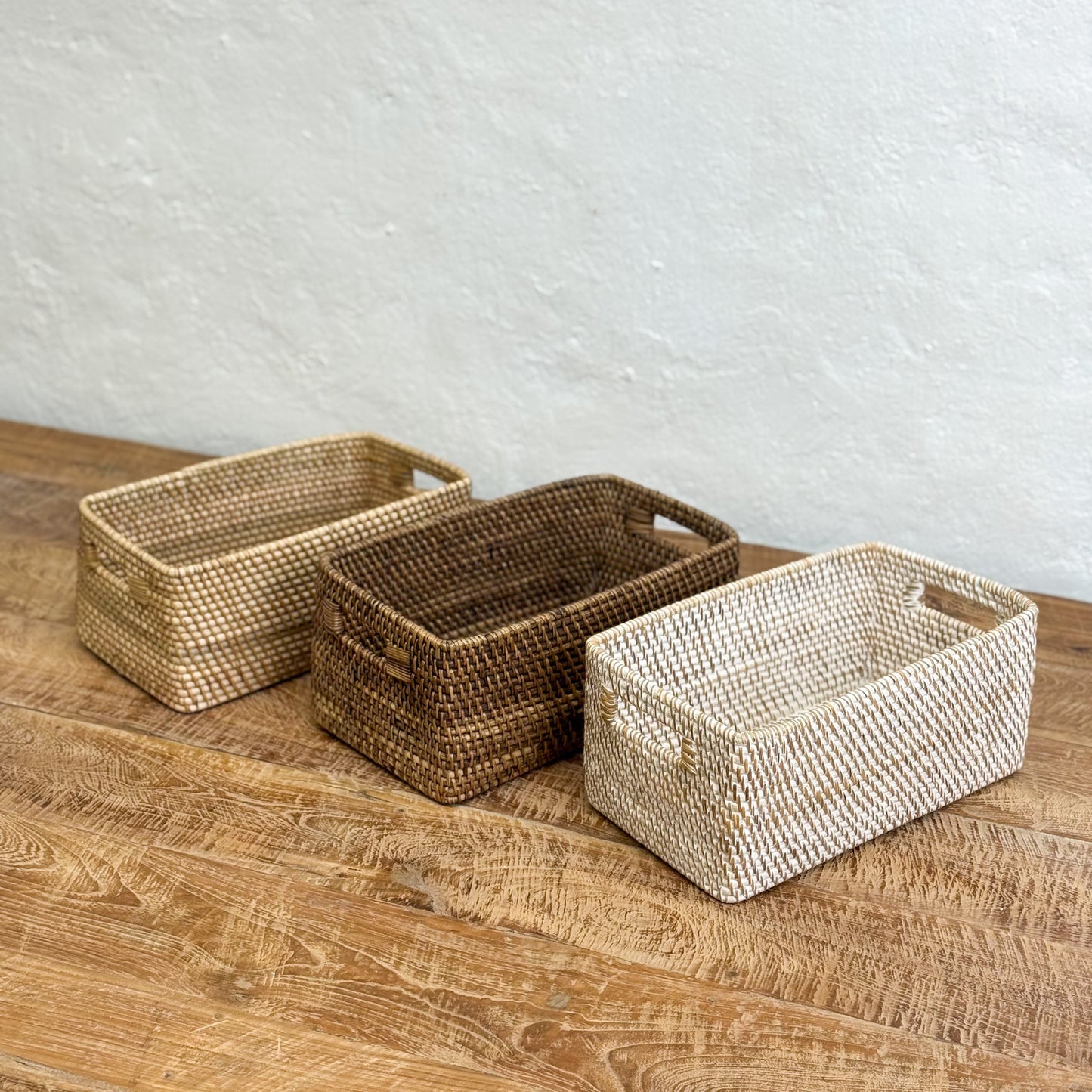 Three woven baskets of different sizes on a wooden surface with a white textured wall background.