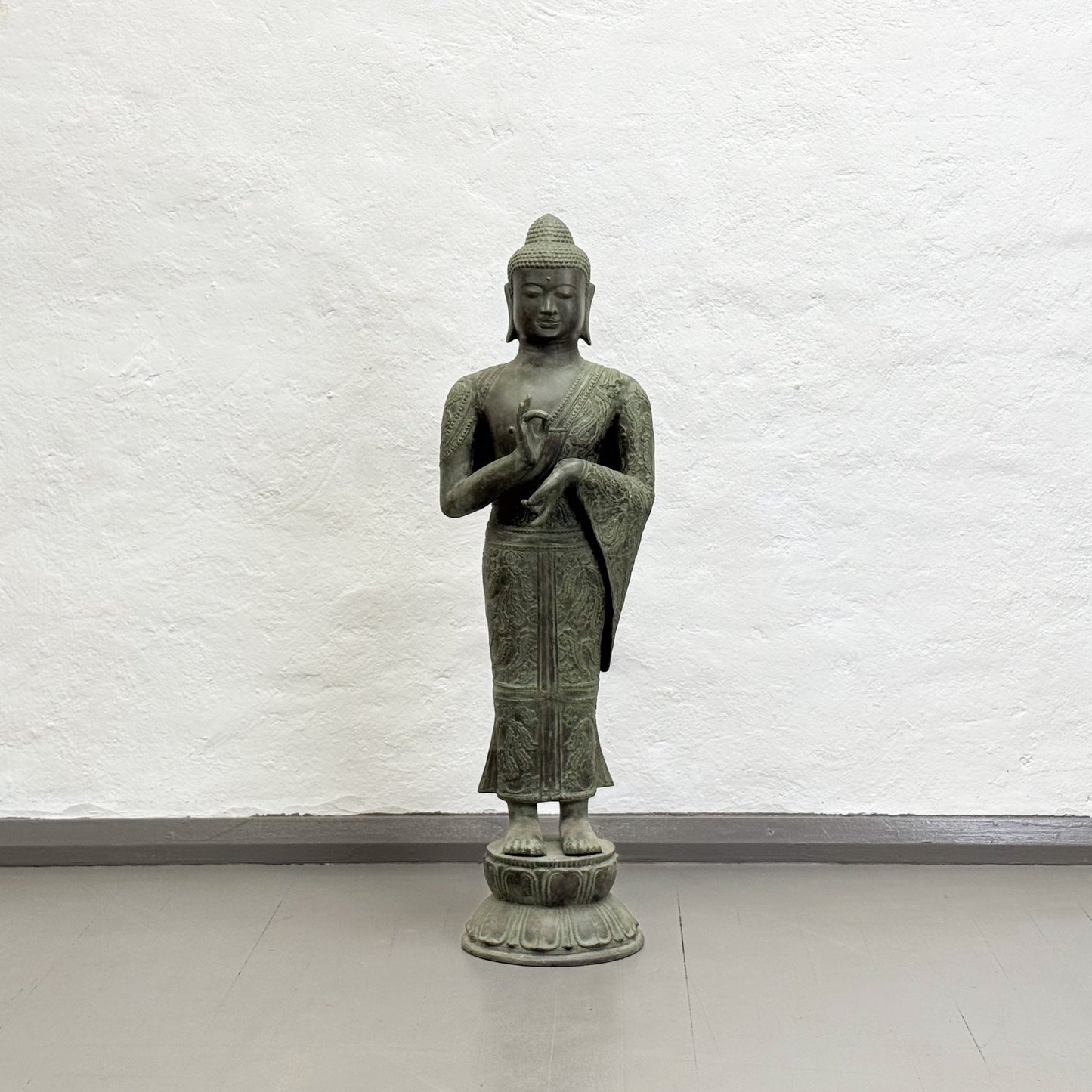 Bronze statue of a standing Buddha against a white brick wall.