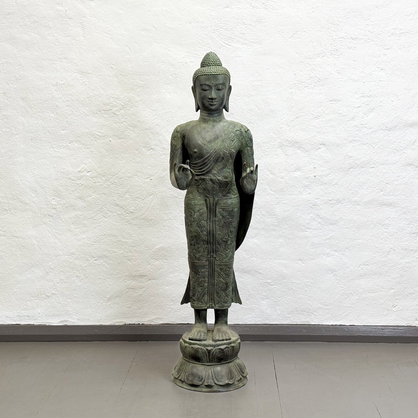Bronze statue of a standing Buddha against a white textured wall.