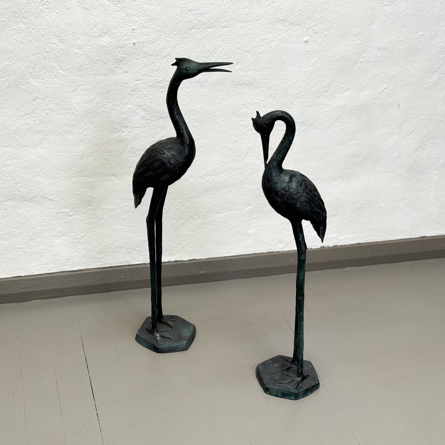 Two bronze cranes on stands against a white wall.