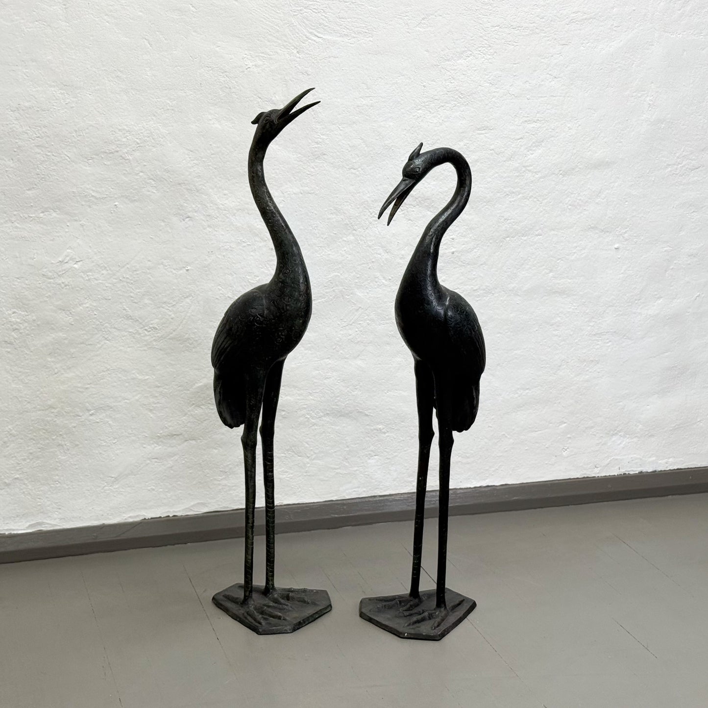 Two black metal crane sculptures on a light gray floor against a white textured wall.