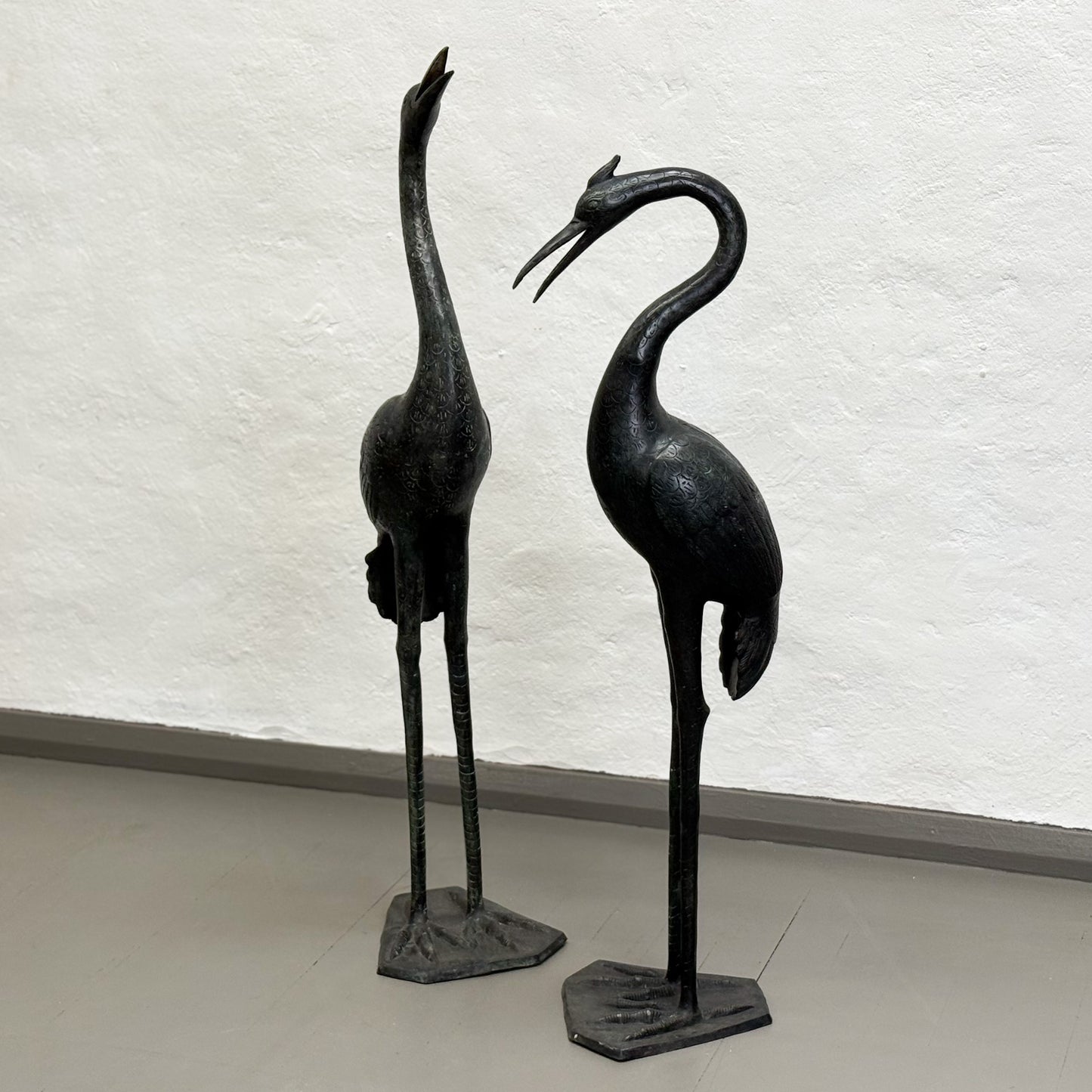 Two black metal crane sculptures on a light gray floor with a white textured wall background.