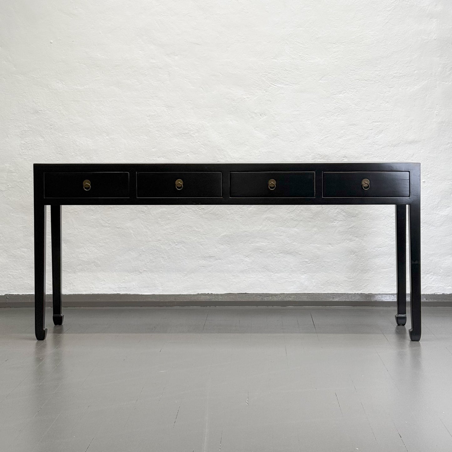 A black wooden console table with four drawers and hoofed legs, placed against a plain wall on a grey floor.