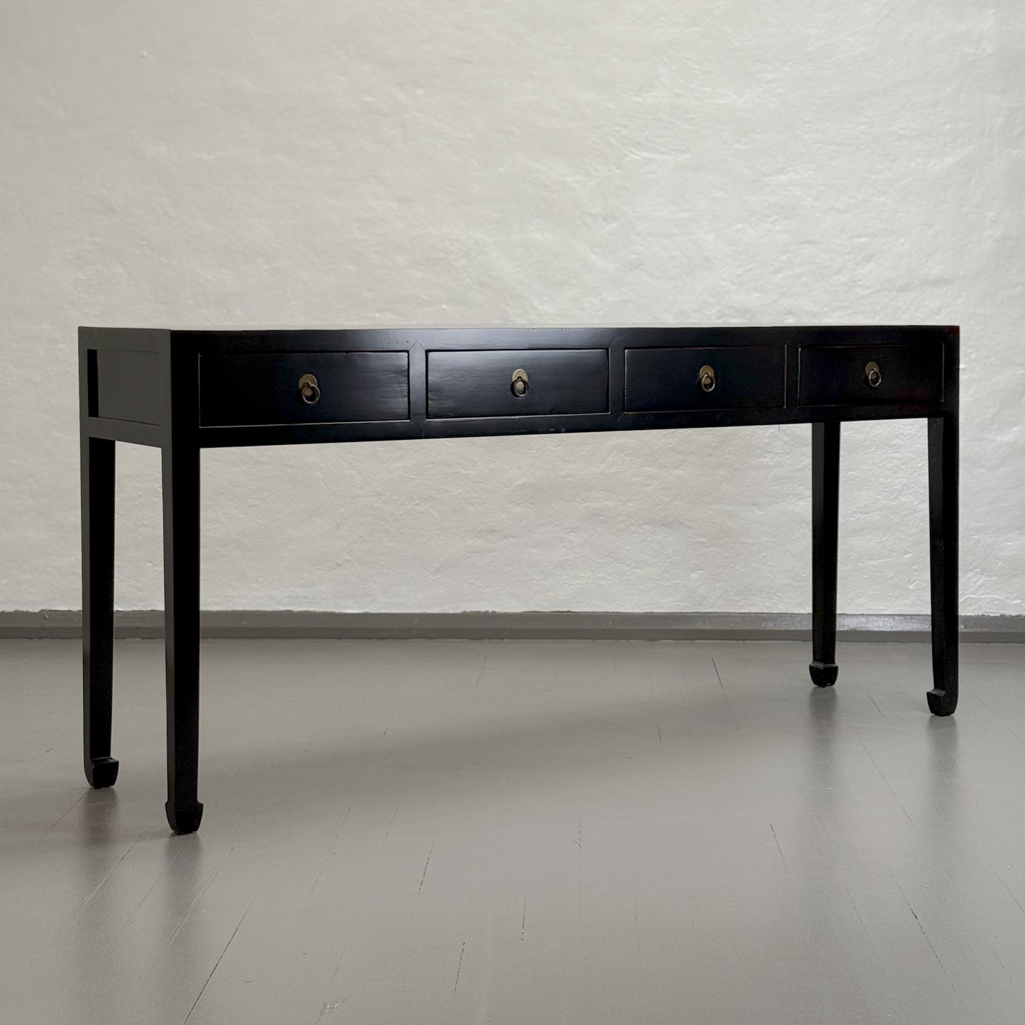 A black wooden console table with four drawers and hoofed legs, placed against a plain wall on a grey floor.