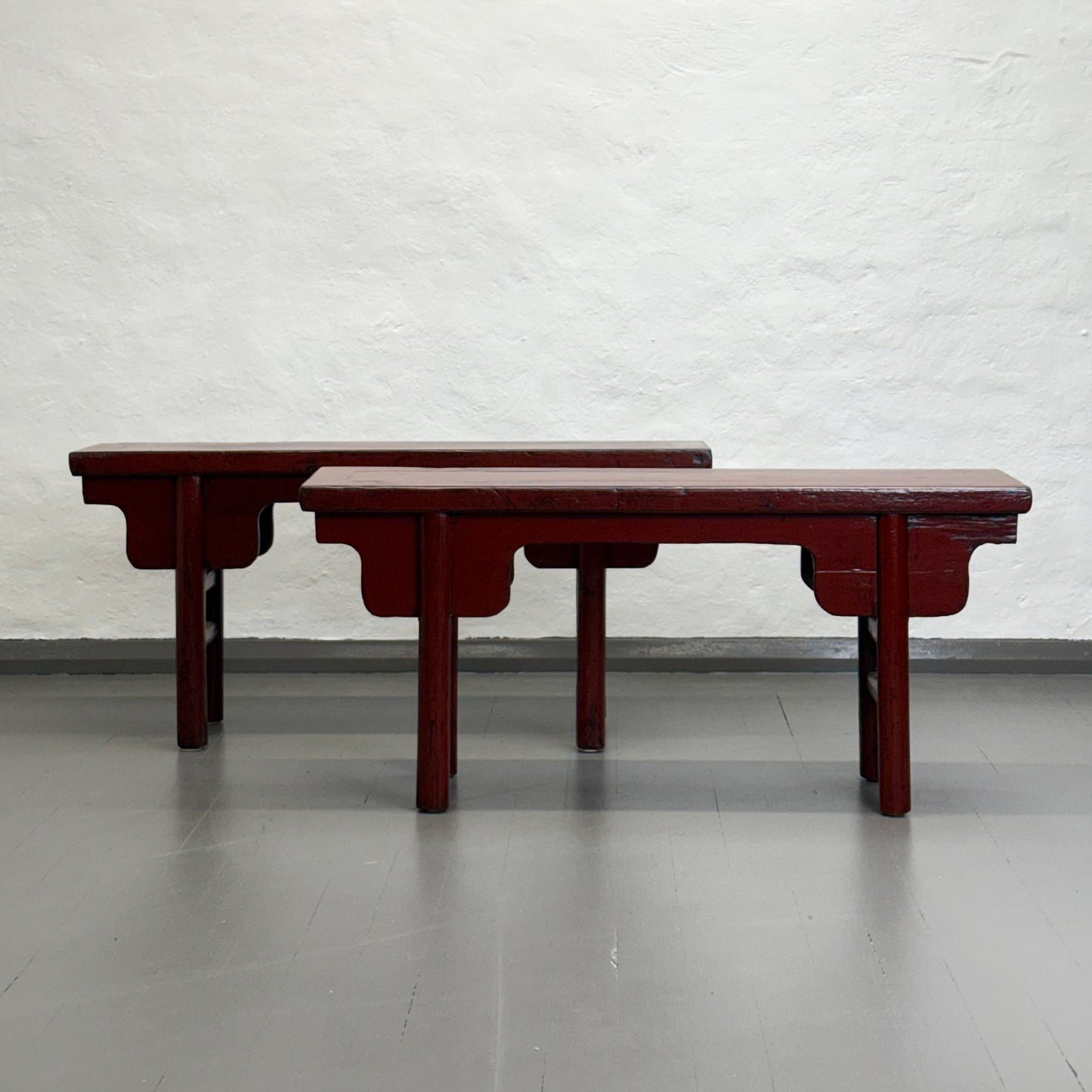 An antique red lacquer bench with a rectangular top and wooden legs, situated against a plain wall on a grey floor.