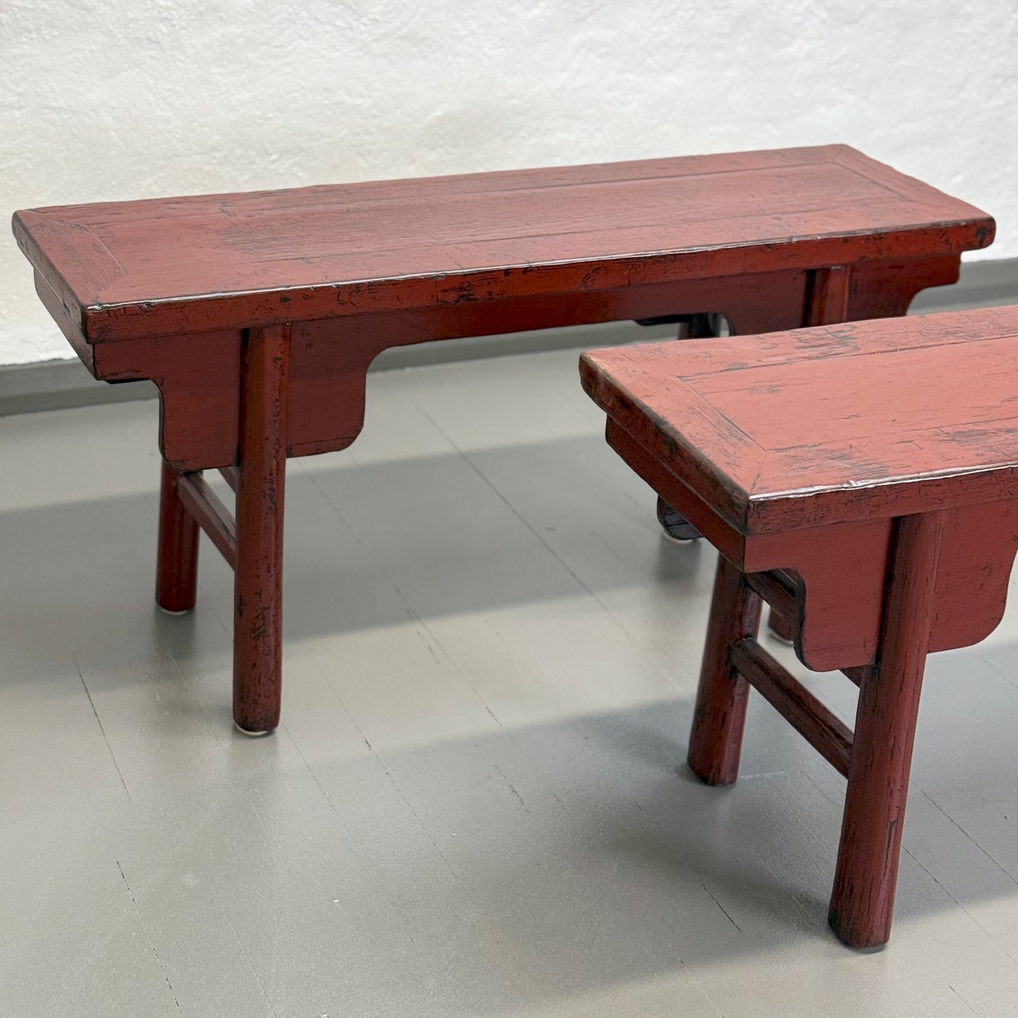 An antique red lacquer bench with a rectangular top and wooden legs, situated against a plain wall on a grey floor.
