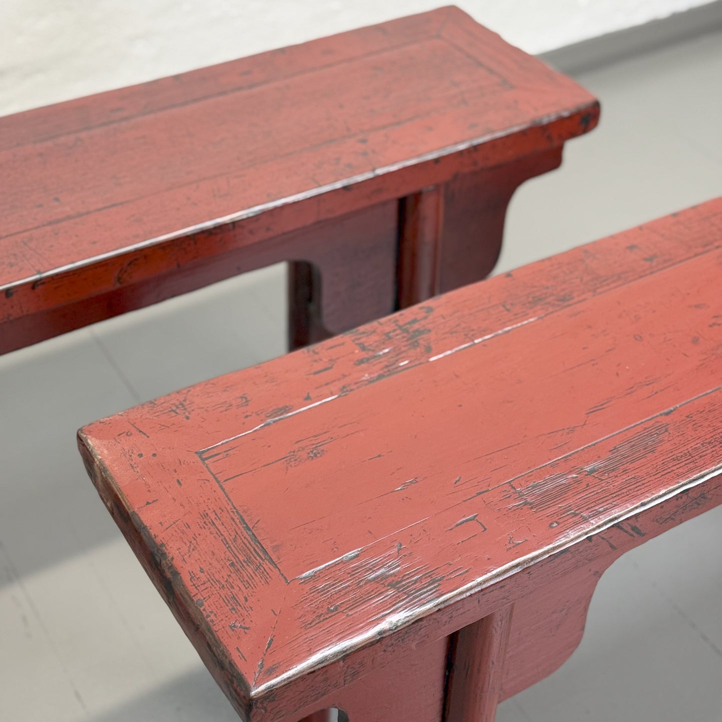 An antique red lacquer bench with a rectangular top and wooden legs, situated against a plain wall on a grey floor.