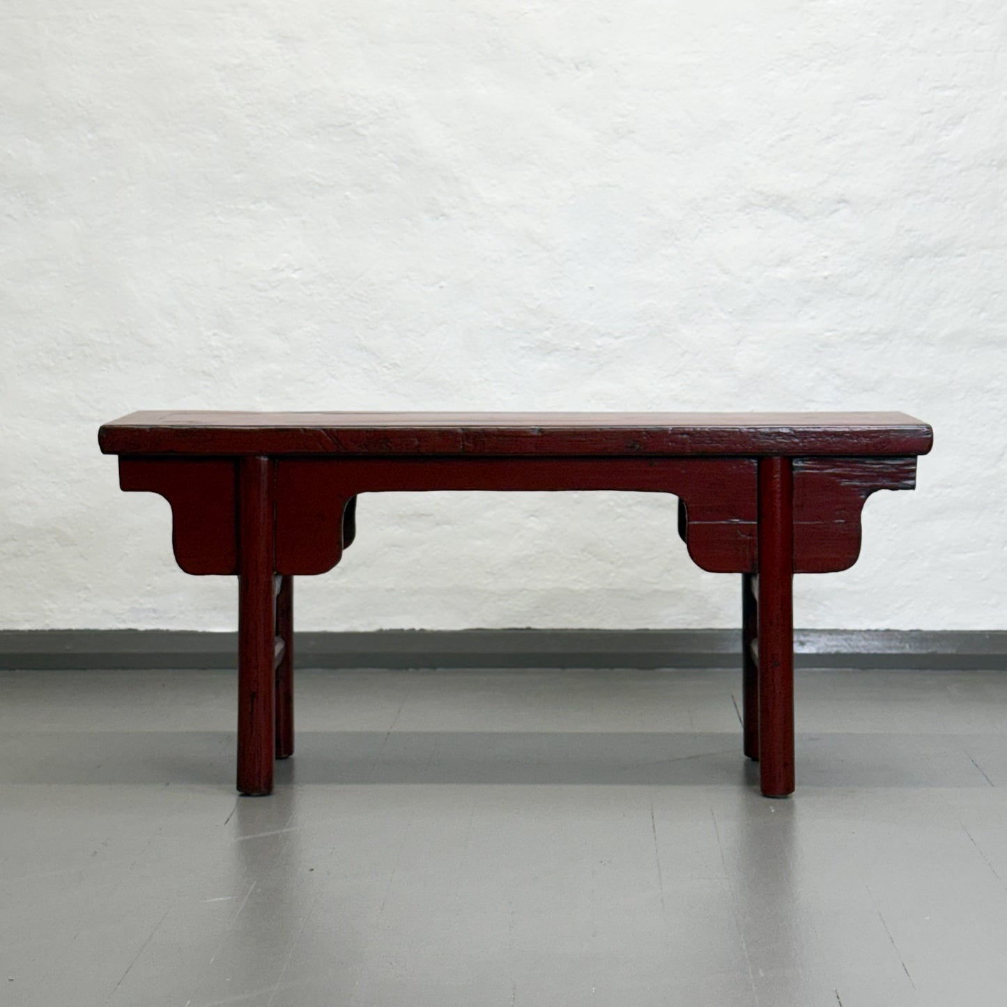 An antique red lacquer bench with a rectangular top and wooden legs, situated against a plain wall on a grey floor.