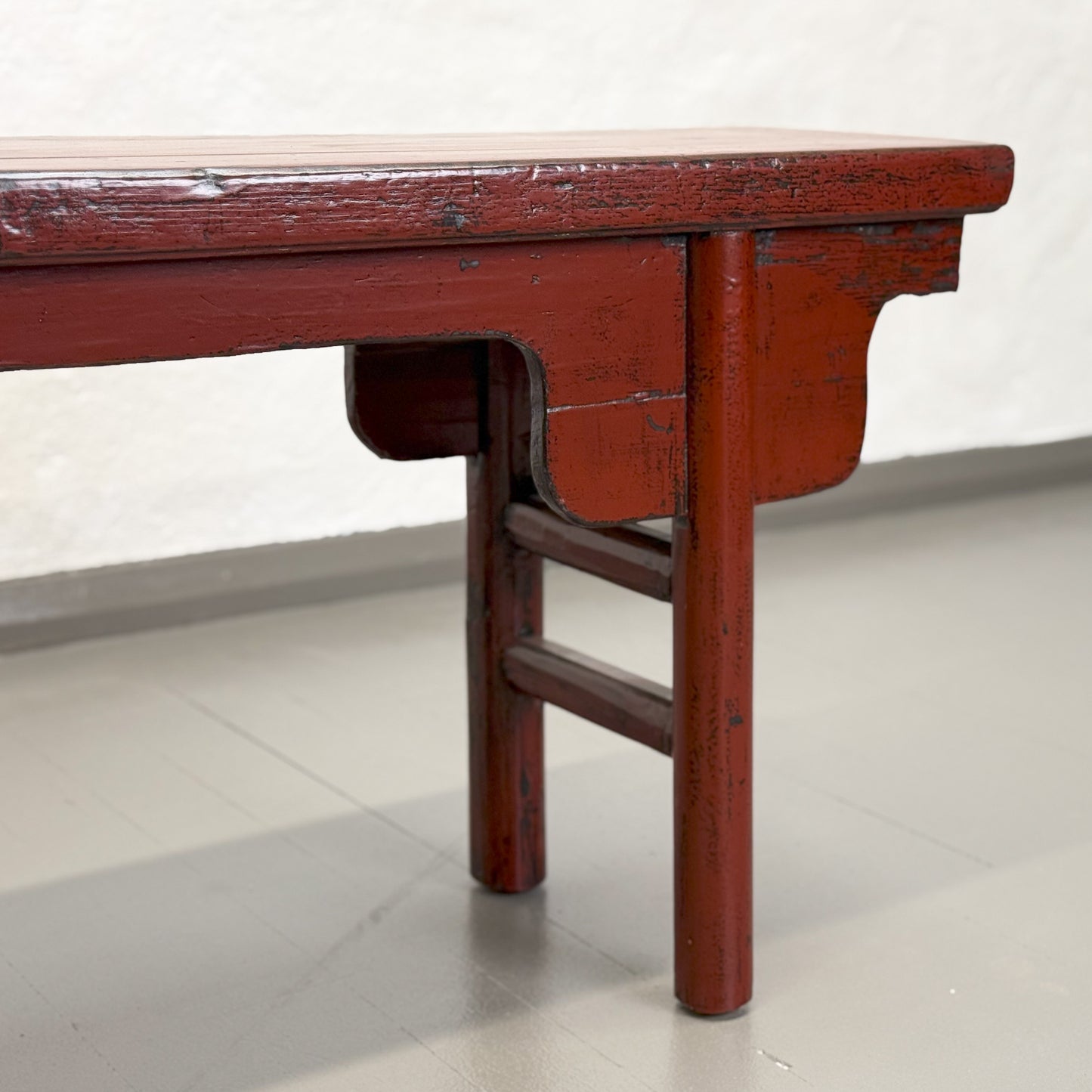 An antique red lacquer bench with a rectangular top and wooden legs, situated against a plain wall on a grey floor.