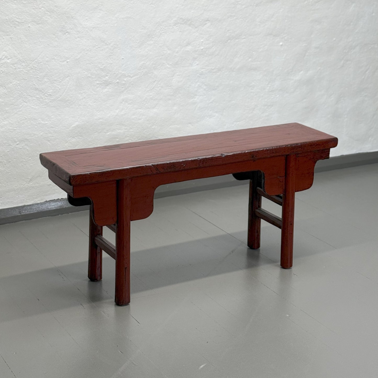 An antique red lacquer bench with a rectangular top and wooden legs, situated against a plain wall on a grey floor.