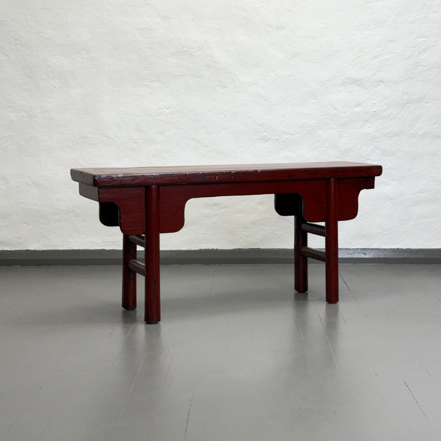 An antique red lacquer bench with a rectangular top and wooden legs, situated against a plain wall on a grey floor.
