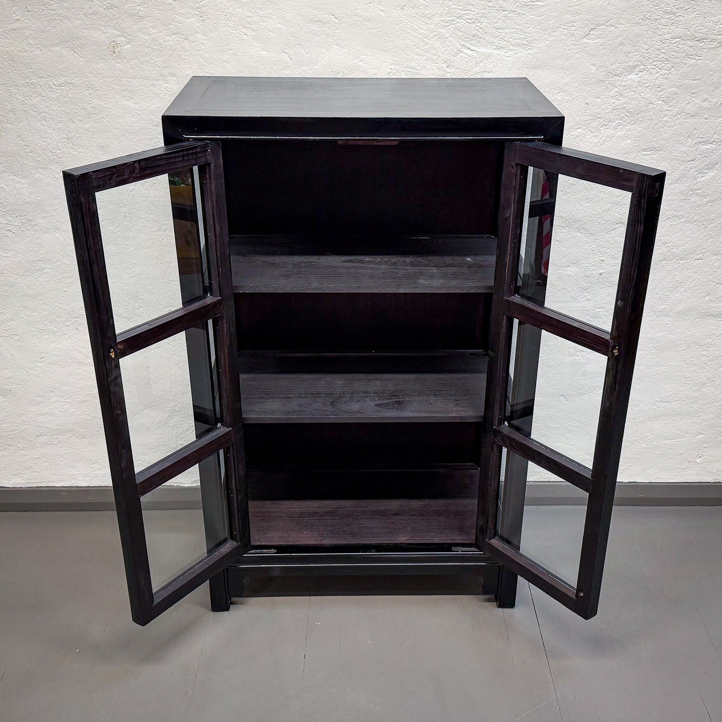 Dark wooden cabinet with glass doors on a light gray floor and white textured wall background