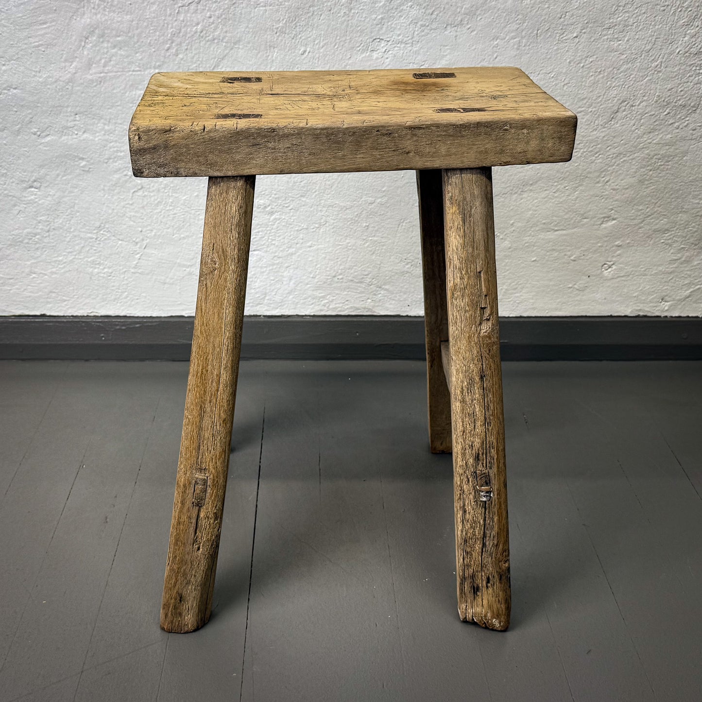Wooden stool with a rustic design on a gray floor against a white wall.