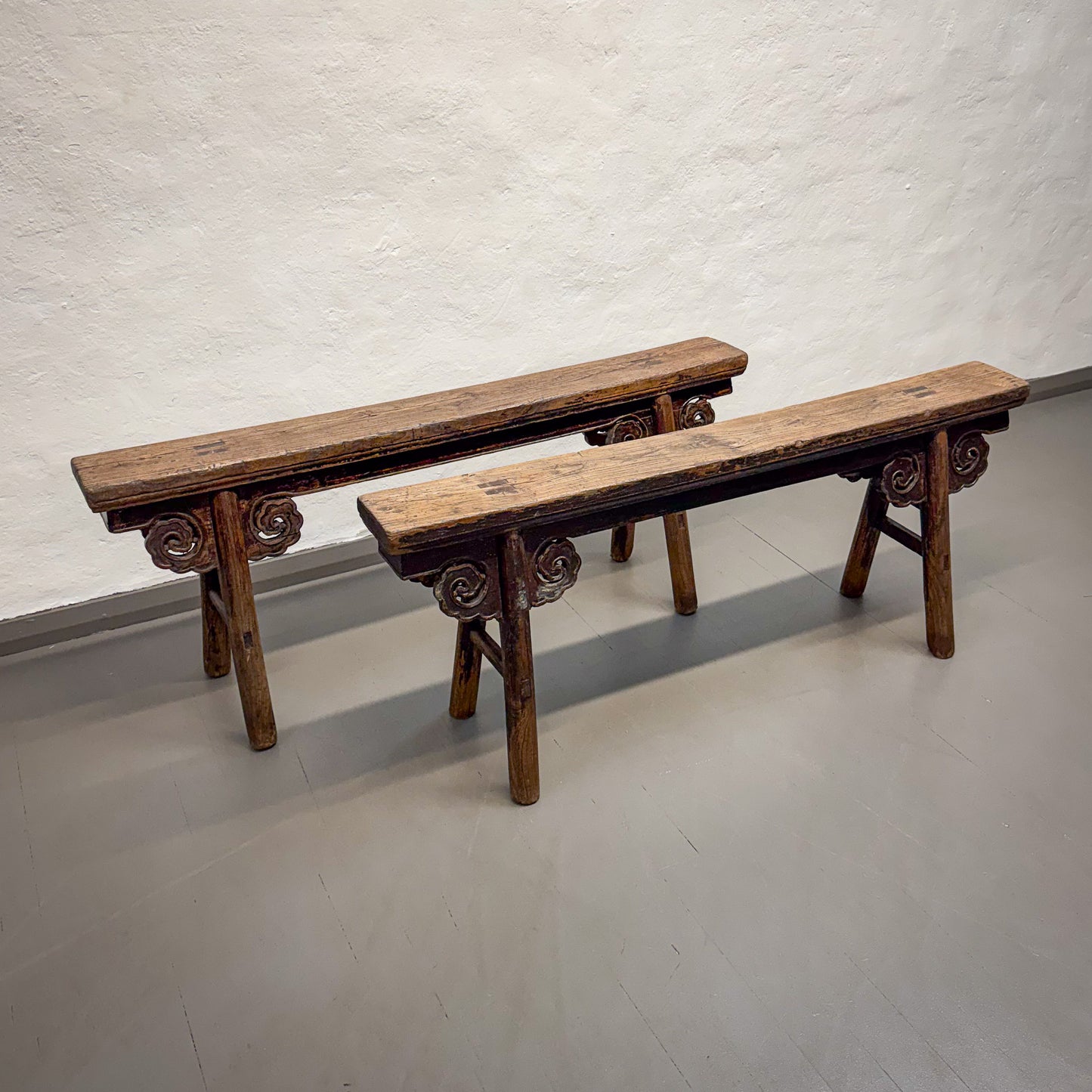 Two wooden benches with decorative carvings on a plain floor and wall.