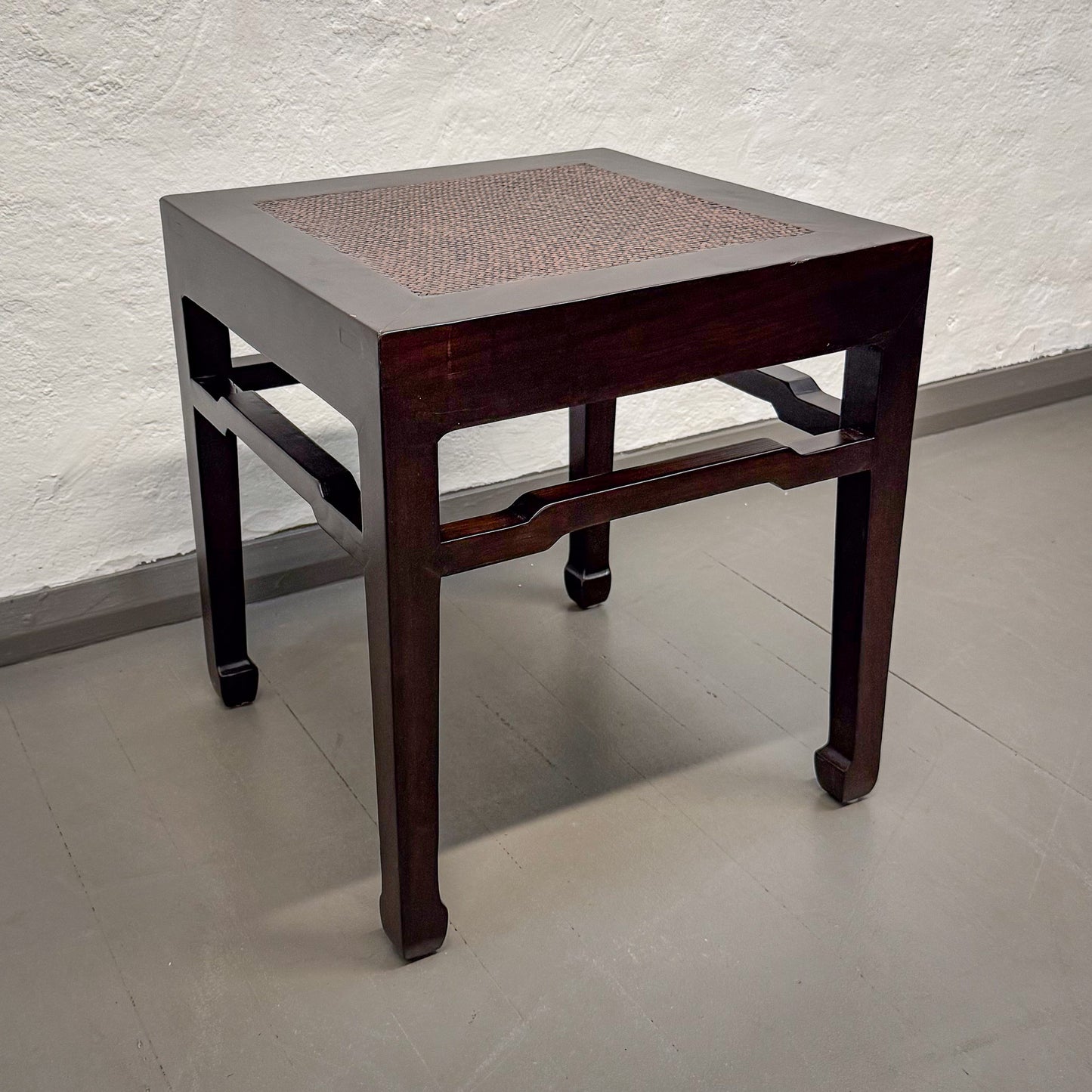 Small square table with a woven top on a plain floor against a textured wall.