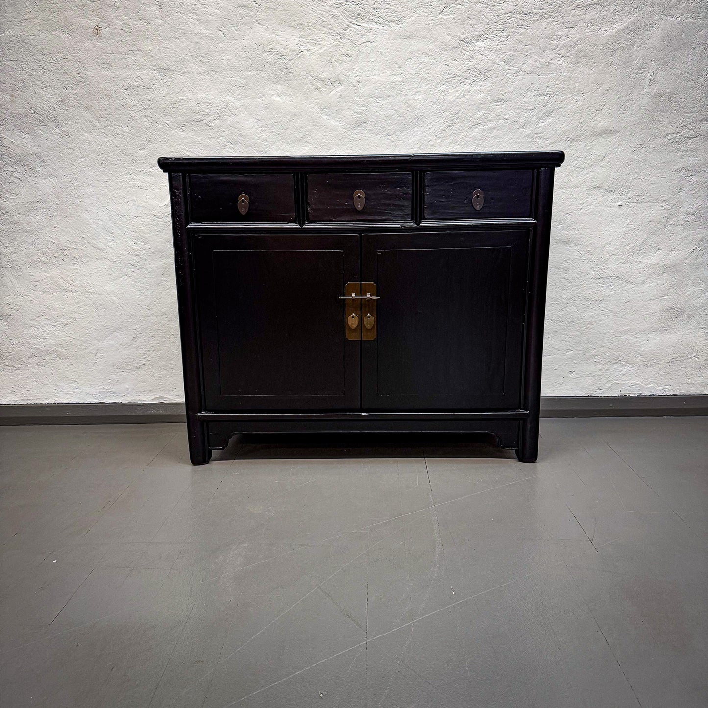 Black wooden cabinet with two doors and two drawers on a gray floor against a textured white wall.