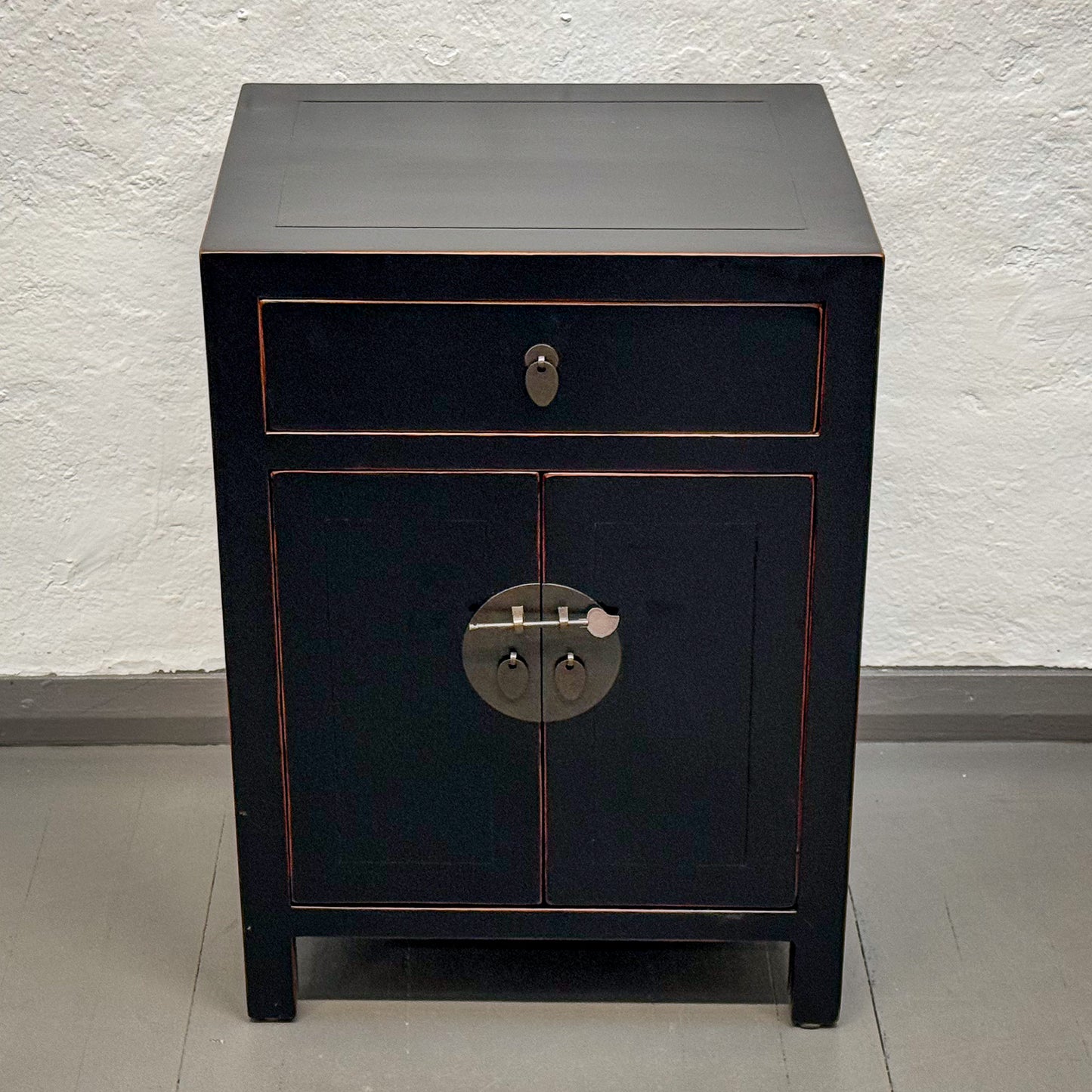 Black wooden cabinet with a single drawer and circular handle on a white textured wall background