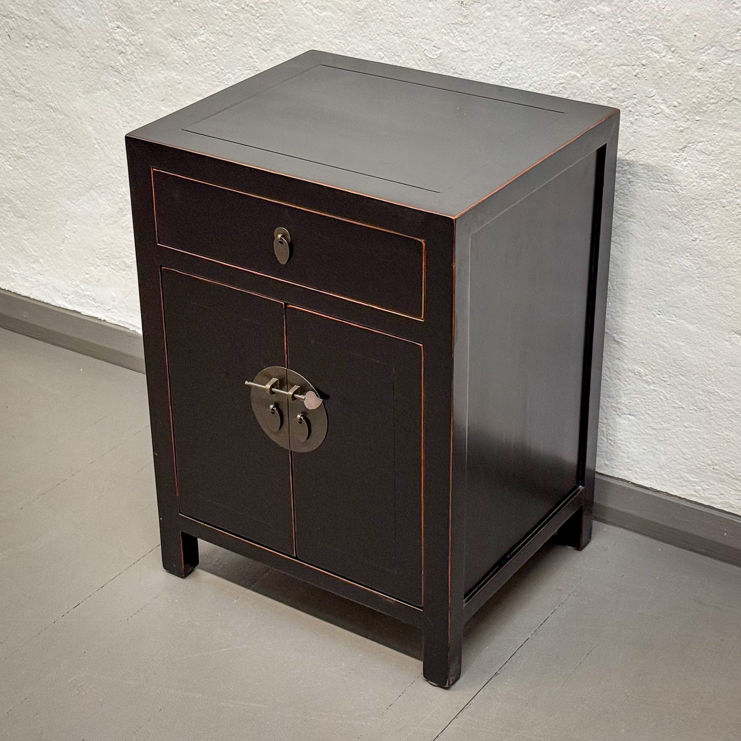 Dark wooden side table with a single drawer and decorative lock against a light wall.