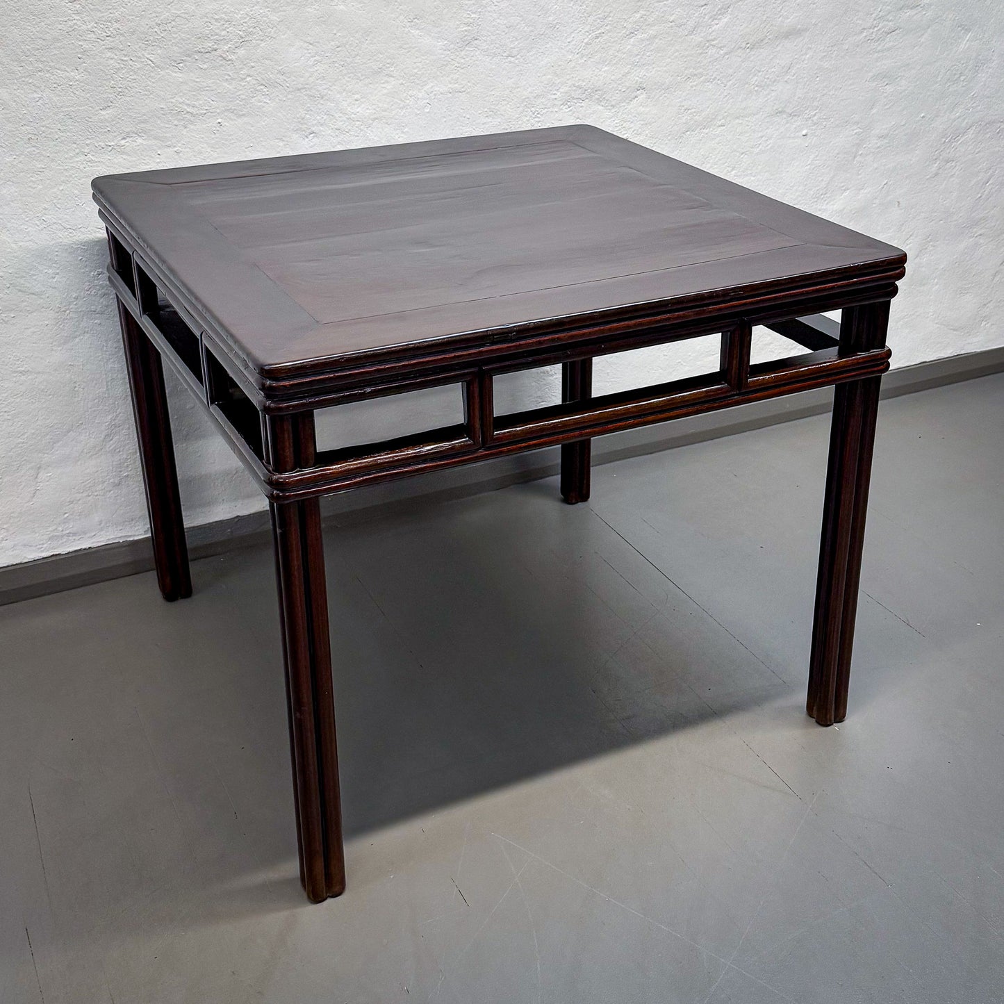 Square wooden table with dark brown finish on a gray floor against a white wall.