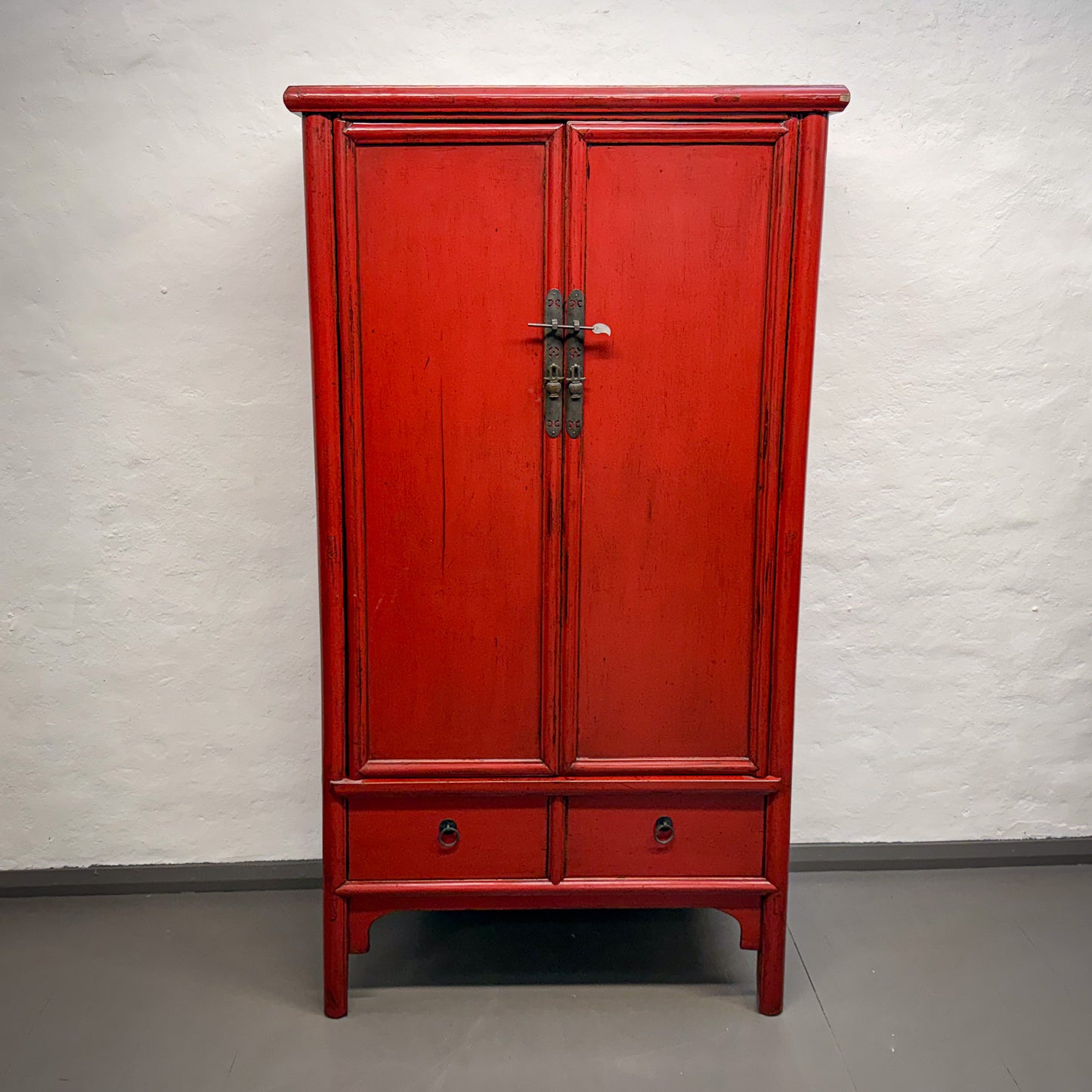 Red wooden cabinet with two doors and a drawer on a gray floor against a white brick wall.