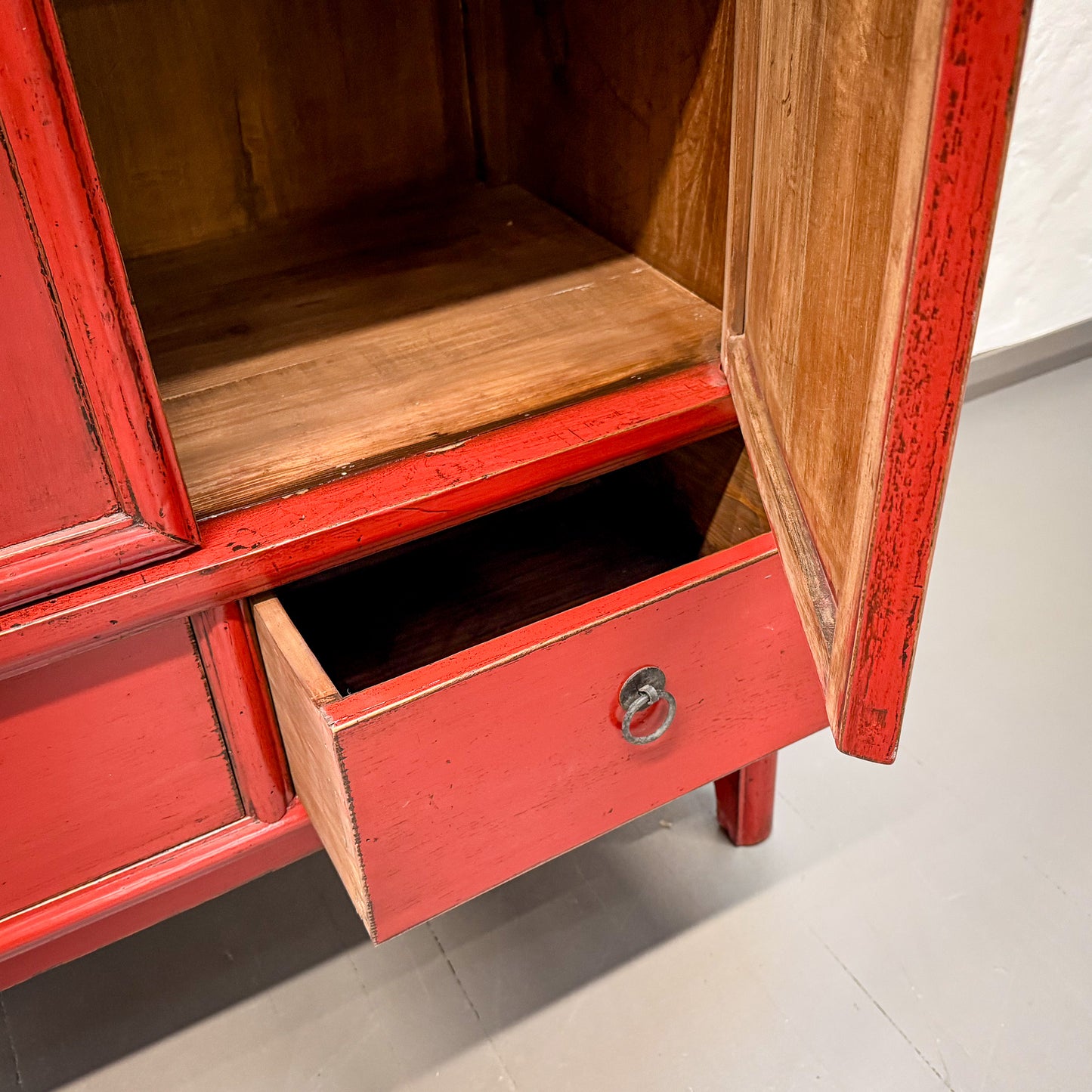 Red wooden cabinet with open door and drawer on a light gray floor.
