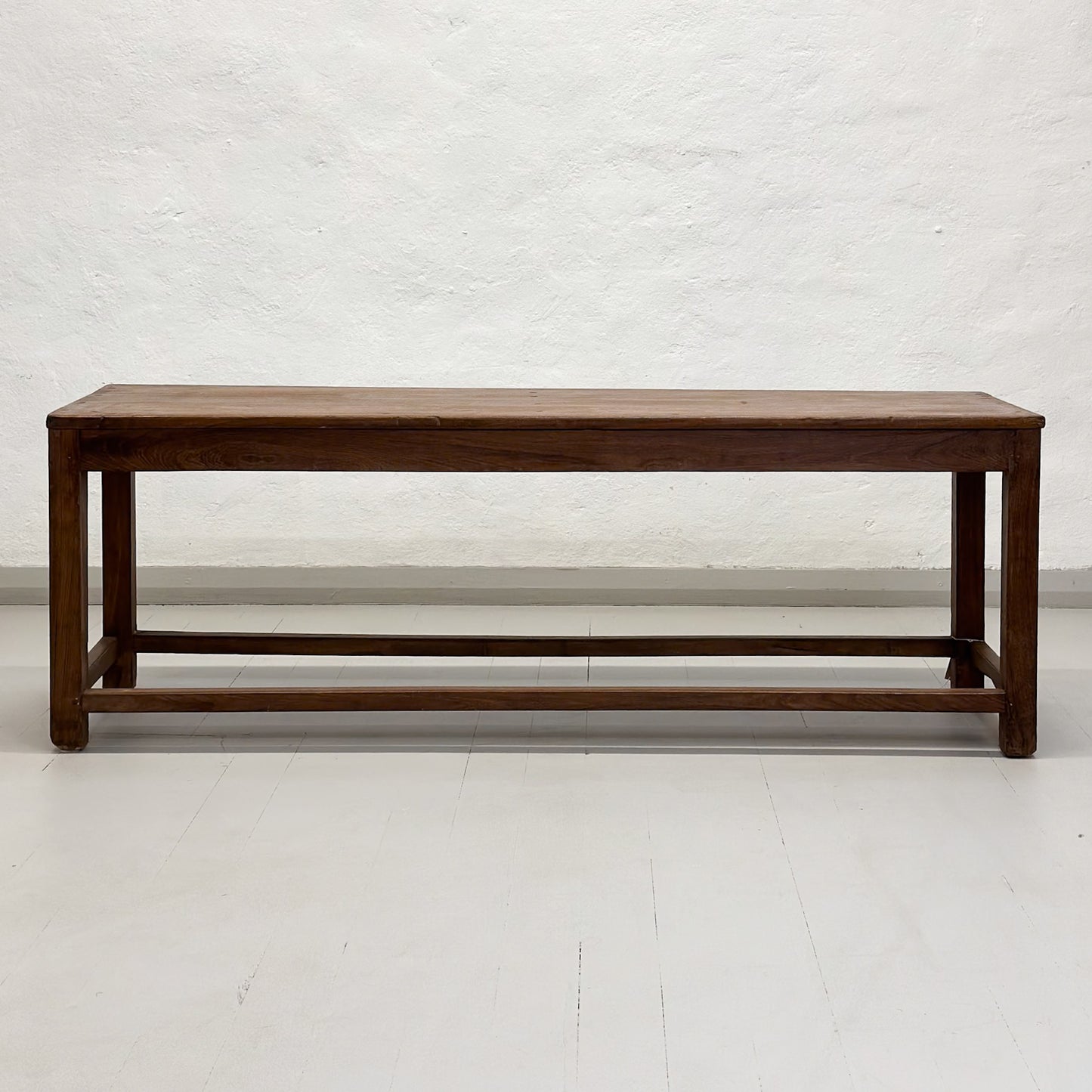 A simple teak wood bench with a slatted backrest and a solid wood seat, placed against a white wall on a concrete floor.