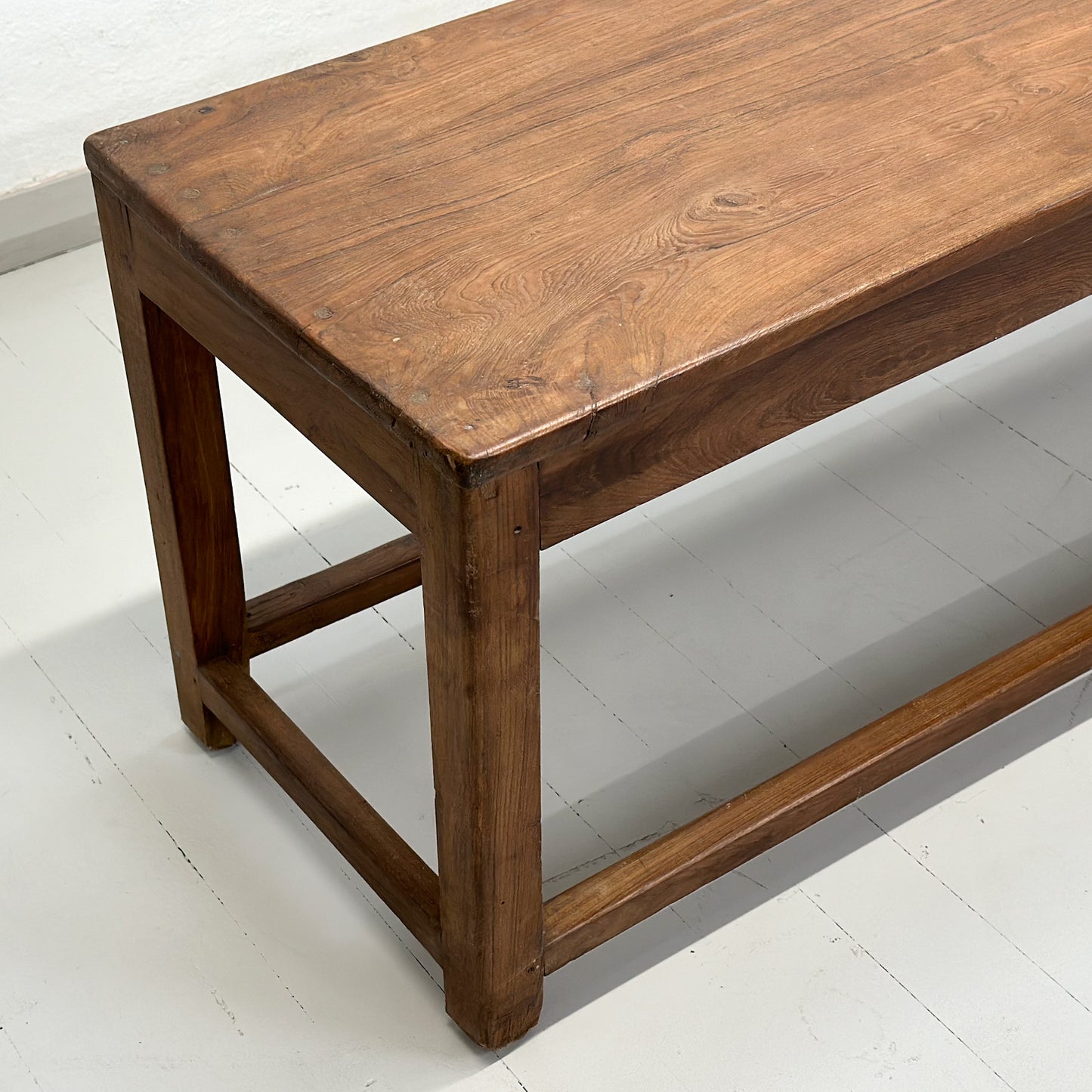 A simple teak wood bench with a slatted backrest and a solid wood seat, placed against a white wall on a concrete floor.
