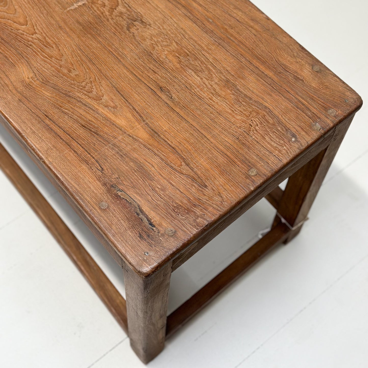 A simple teak wood bench with a slatted backrest and a solid wood seat, placed against a white wall on a concrete floor.
