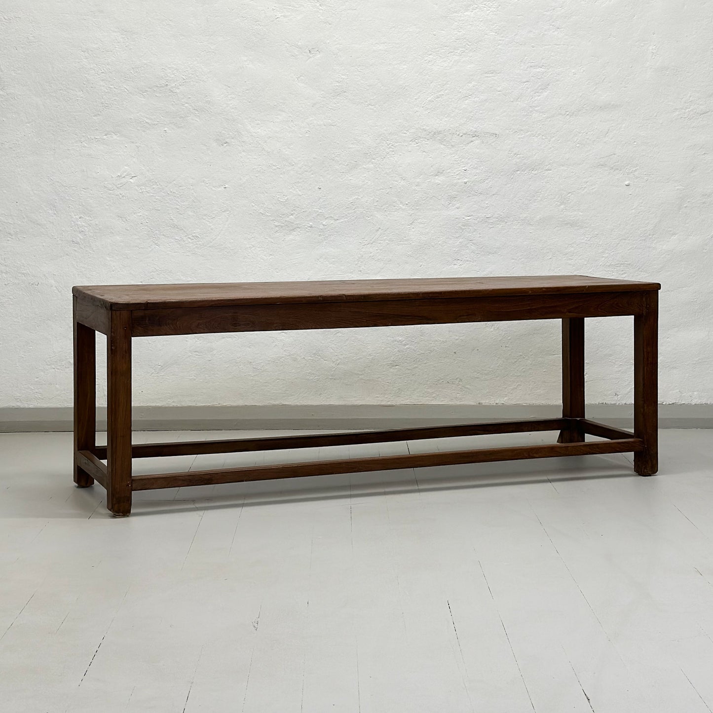 A simple teak wood bench with a slatted backrest and a solid wood seat, placed against a white wall on a concrete floor.