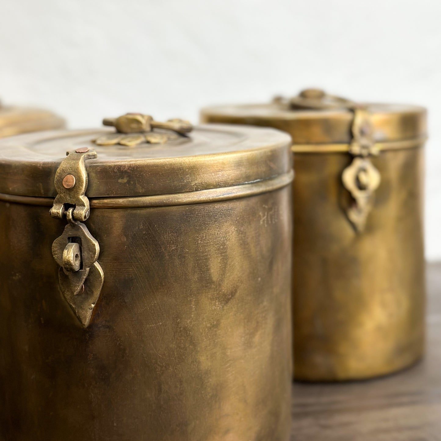 A set of three small brass jars with lids, featuring a decorative design and placed on a wooden surface against a plain wall.