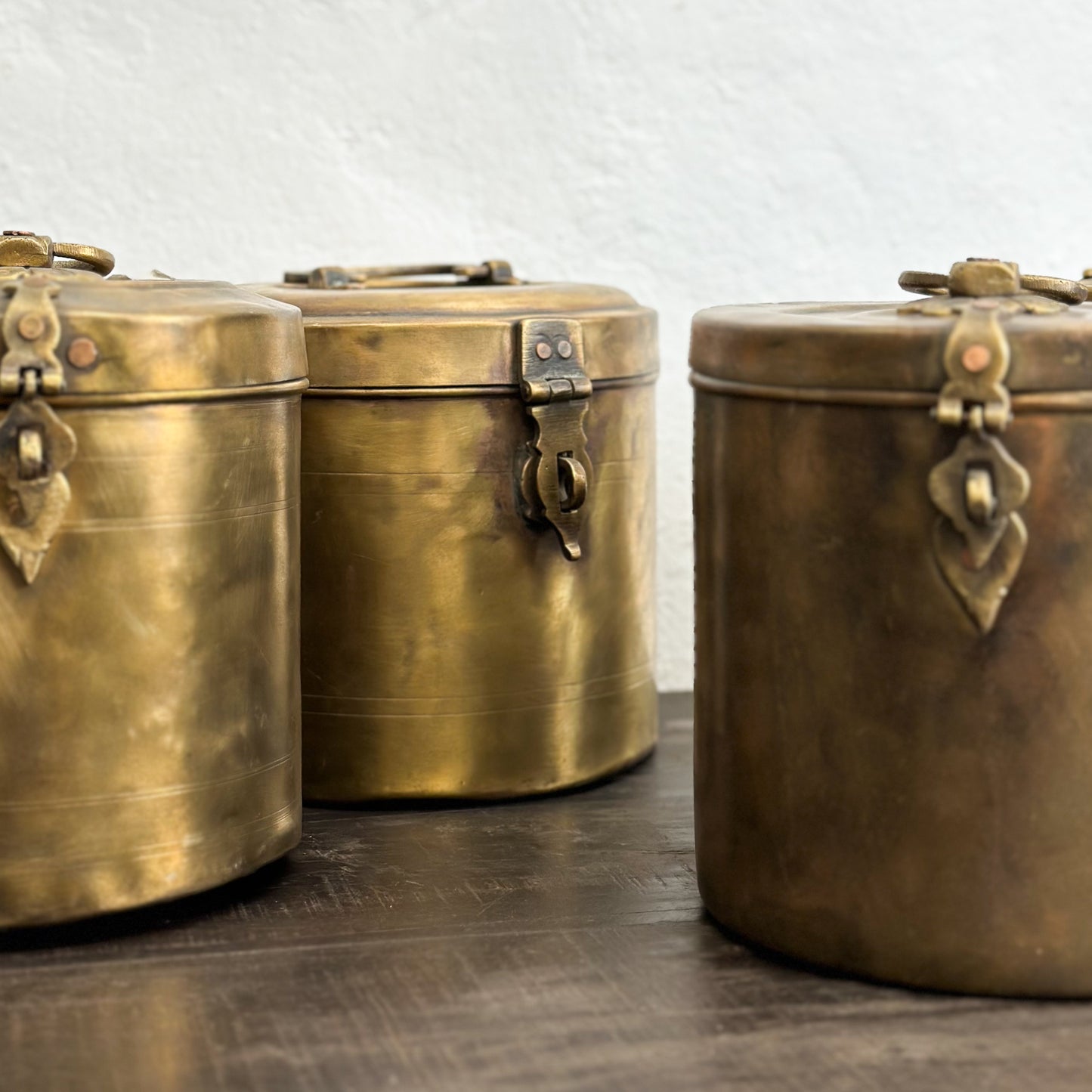 A set of three small brass jars with lids, featuring a decorative design and placed on a wooden surface against a plain wall.