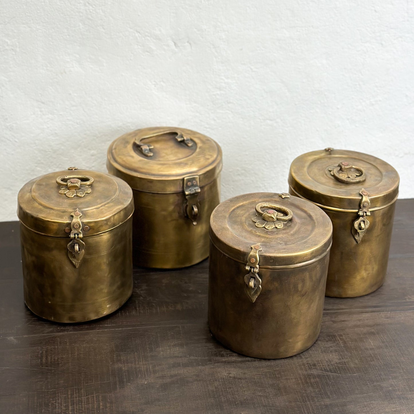 A set of three small brass jars with lids, featuring a decorative design and placed on a wooden surface against a plain wall.