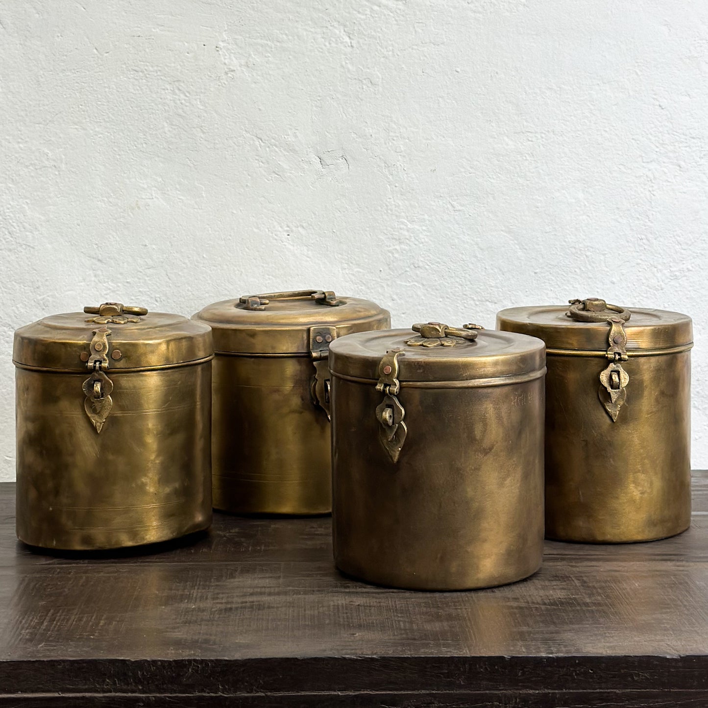 A set of three small brass jars with lids, featuring a decorative design and placed on a wooden surface against a plain wall.