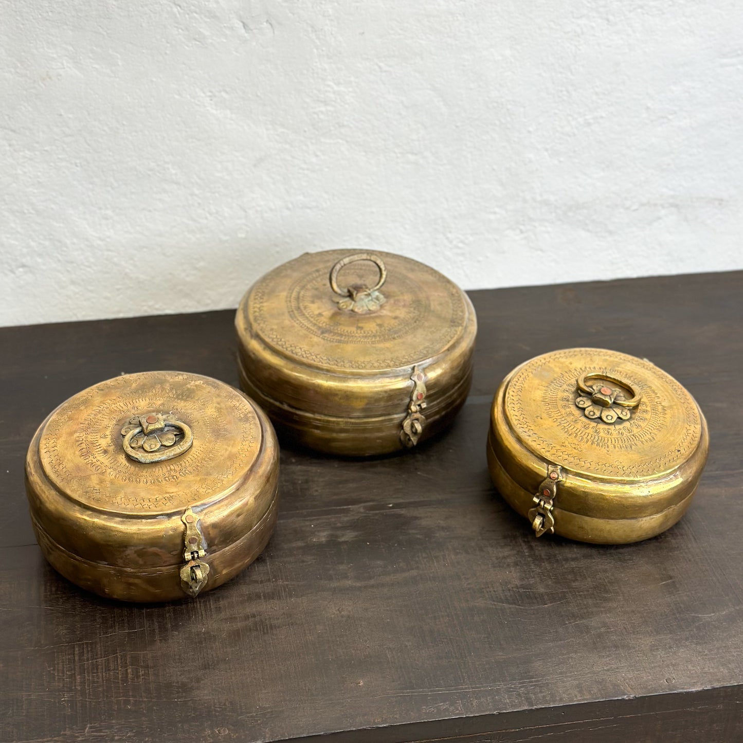 Three brass chapati boxes with lids, placed on a wooden surface against a white wall background.