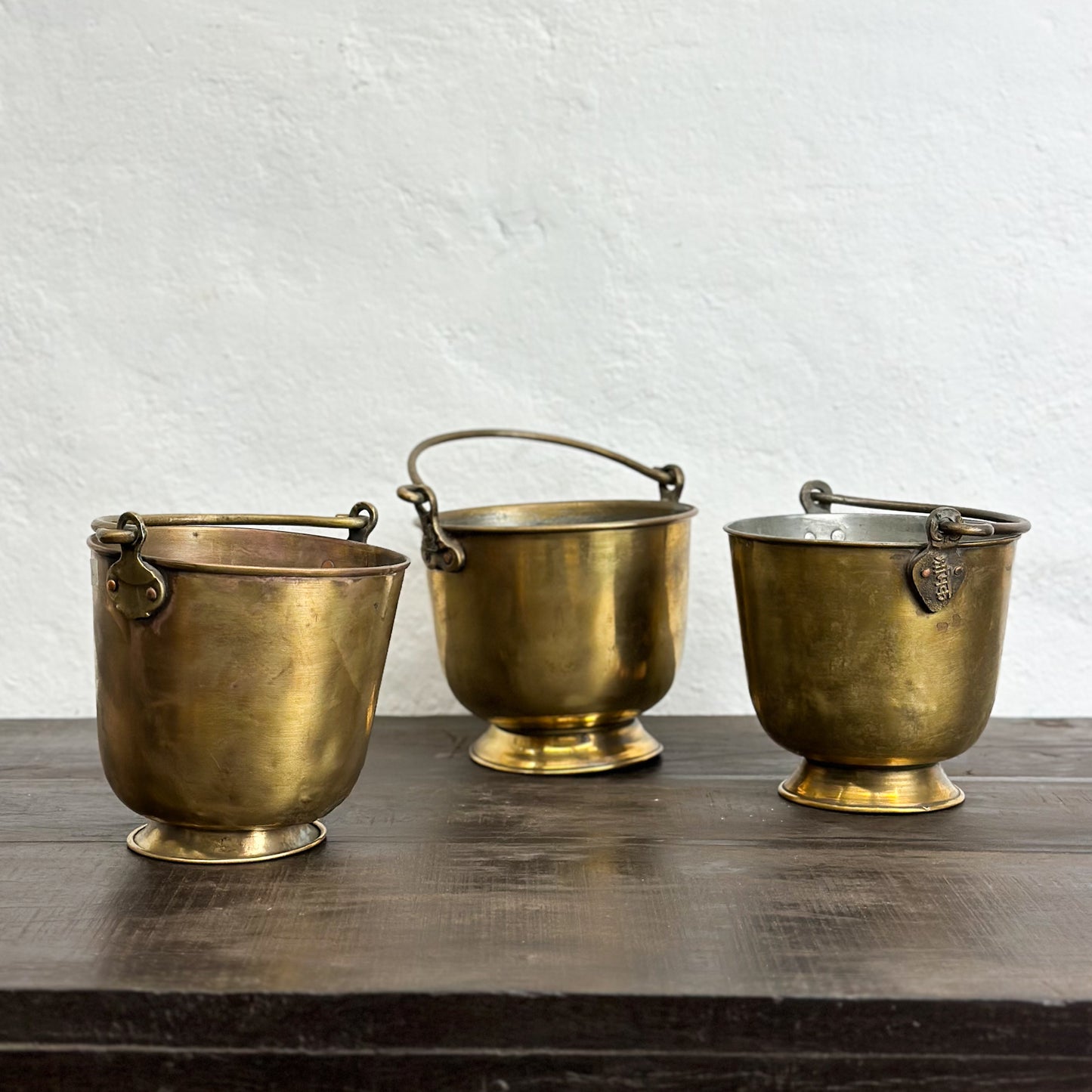 Three small brass buckets with bases, two with handles, placed on a wooden surface against a white wall.
