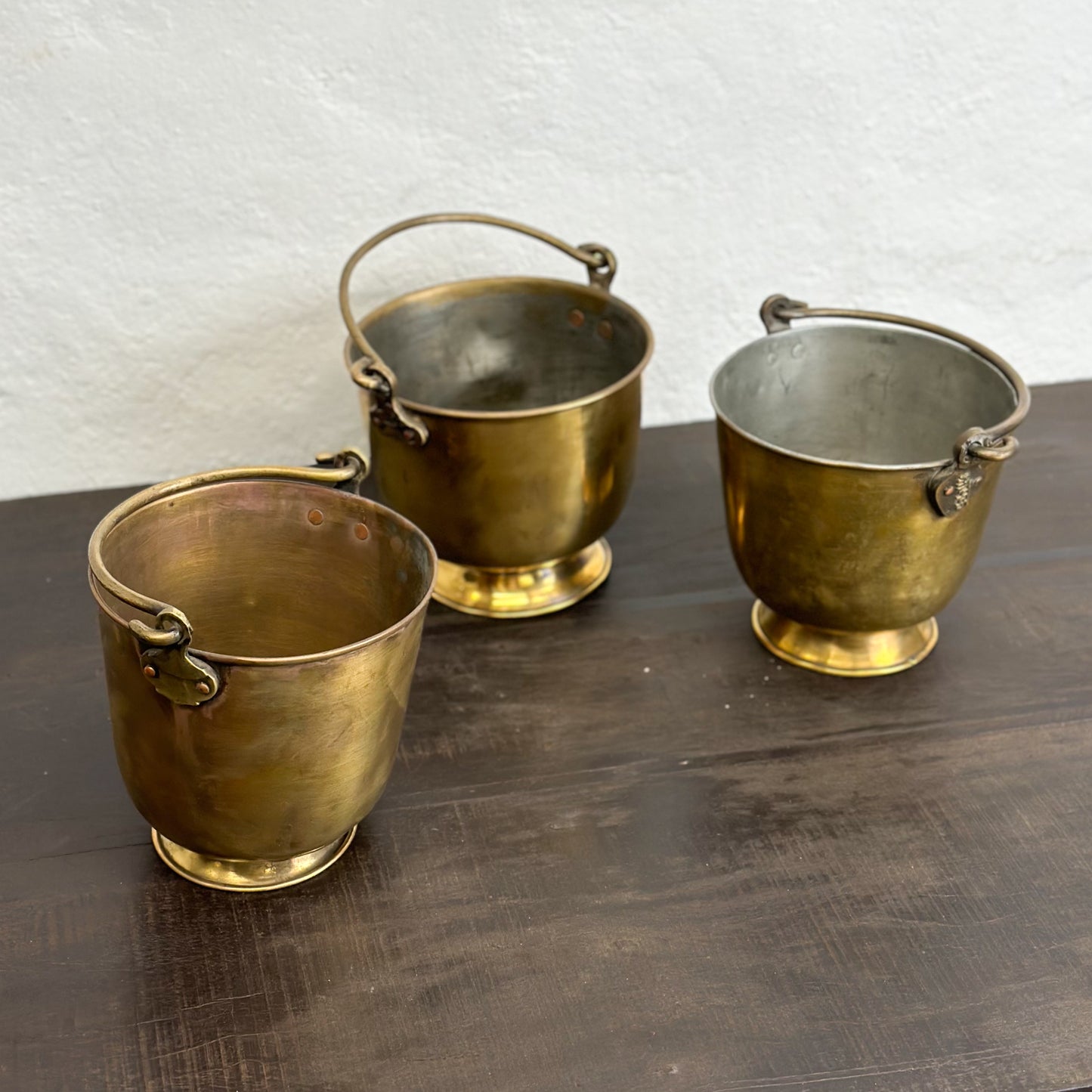 Three small brass buckets with bases, two with handles, placed on a wooden surface against a white wall.