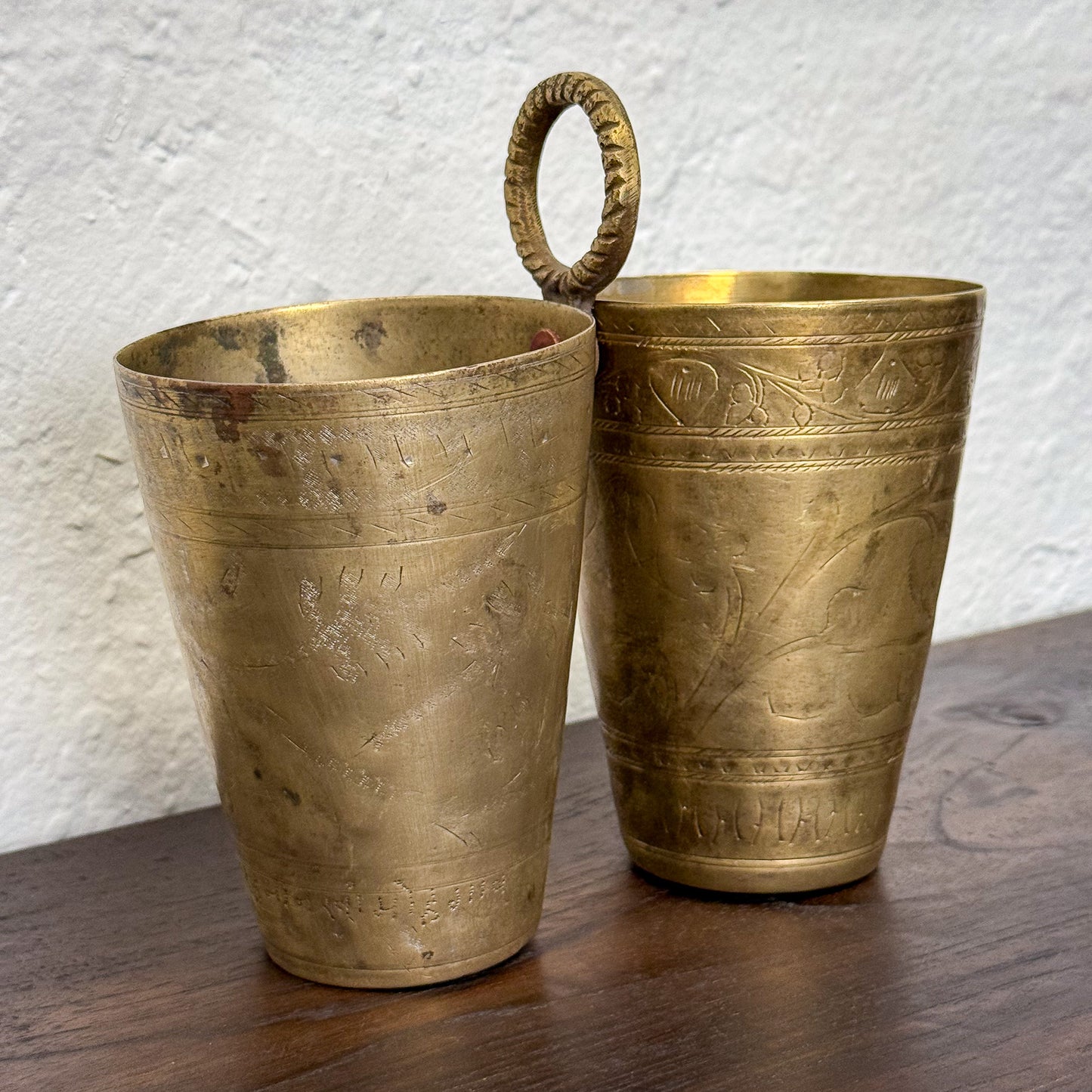 Two brass cups with intricate designs on a wooden surface and textured white background