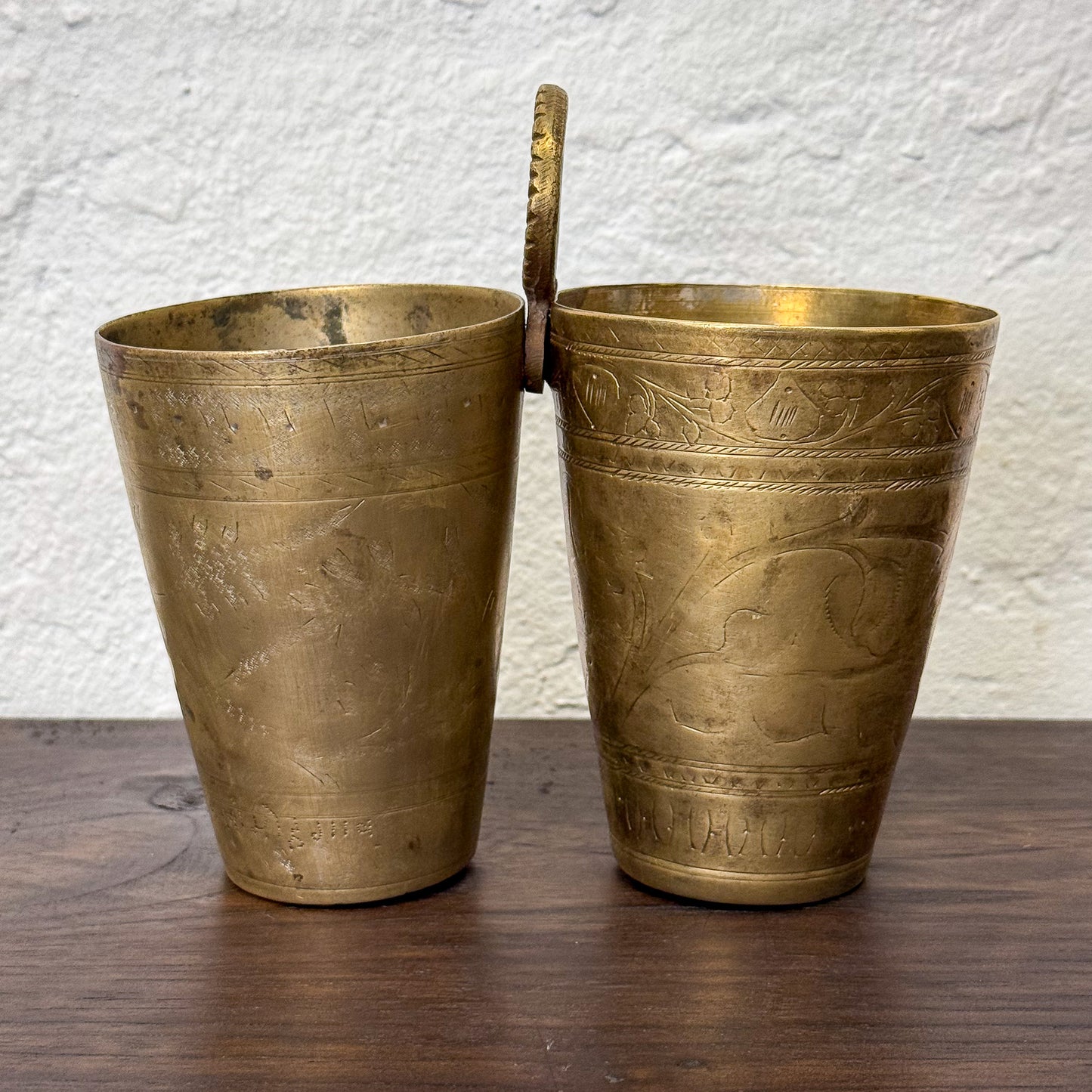 Two brass cups with handles on a wooden surface against a textured white wall.